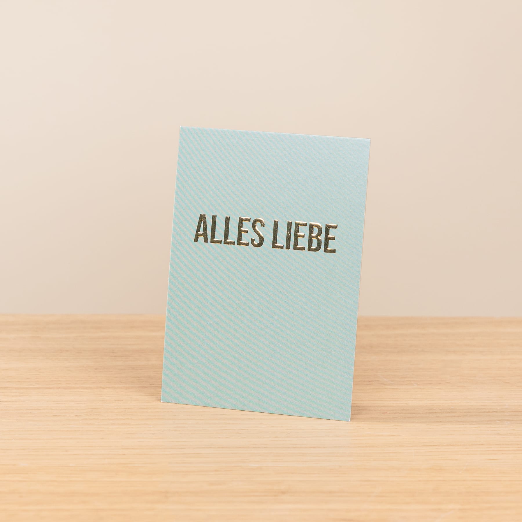 Eine hellblaue Karte mit dem deutschen Spruch „ALLES LIEBE“ liegt auf einer Holzfläche vor beigem Hintergrund.