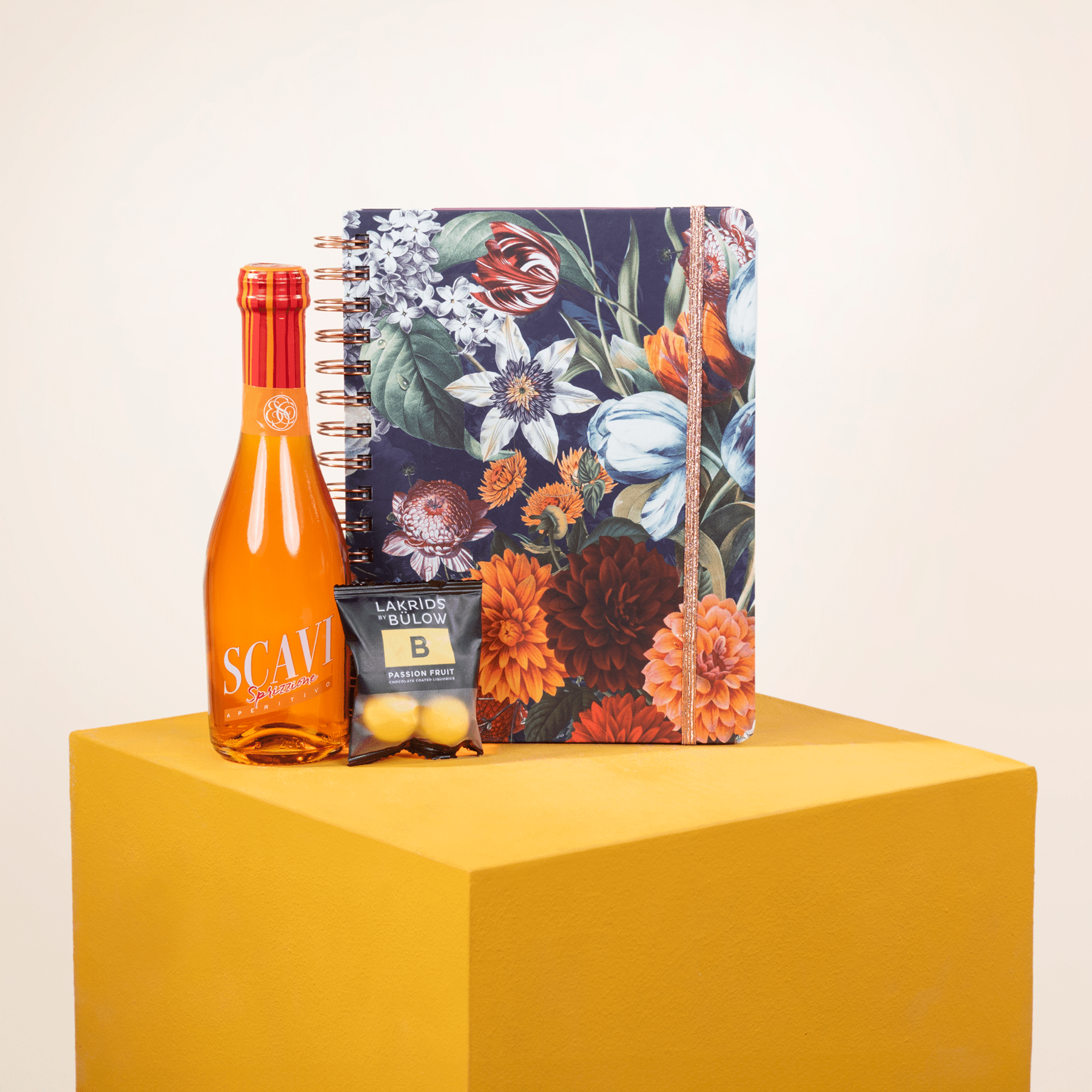 Ein farbenfrohes Notizbuch mit Blumenmuster, eine Flasche Scavi & Ray-Wein und eine kleine Topfpflanze stehen auf einem gelben Würfel und ergeben ein optisch ansprechendes Stillleben-Arrangement.