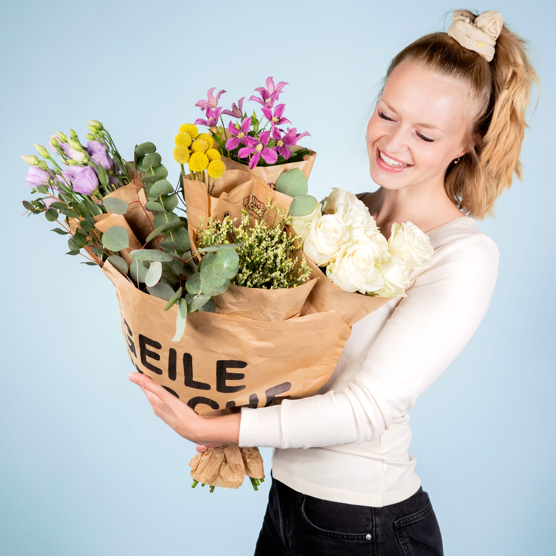 Eine lächelnde junge Frau hält einen großen Strauß bunter Blumen in einer Papiertüte vor einem schlichten blauen Hintergrund.