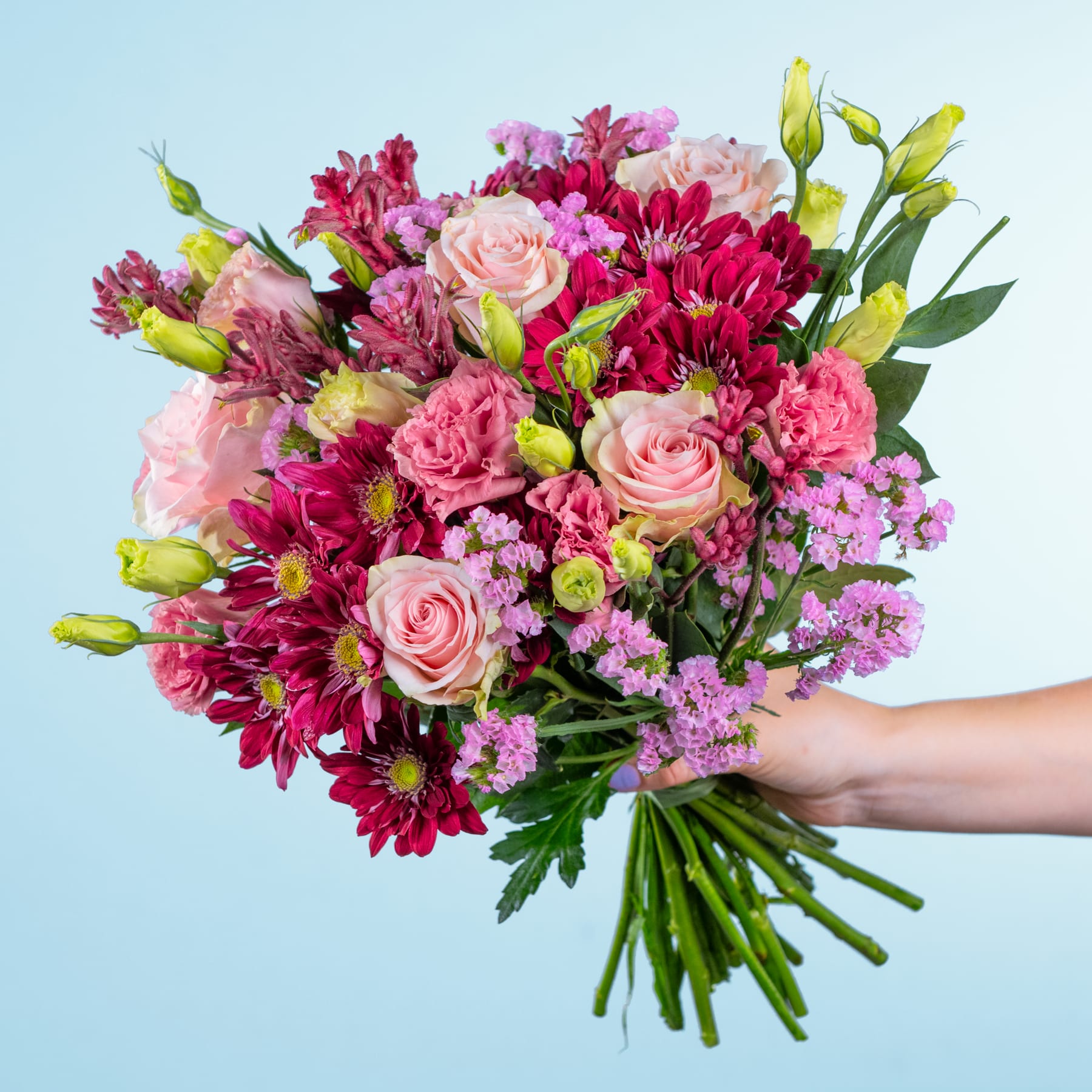 Ein leuchtender Strauß rosa und roter Blumen, darunter Rosen, Nelken und Chrysanthemen, wird vor einem schlichten blauen Hintergrund in der Hand einer Person gehalten.