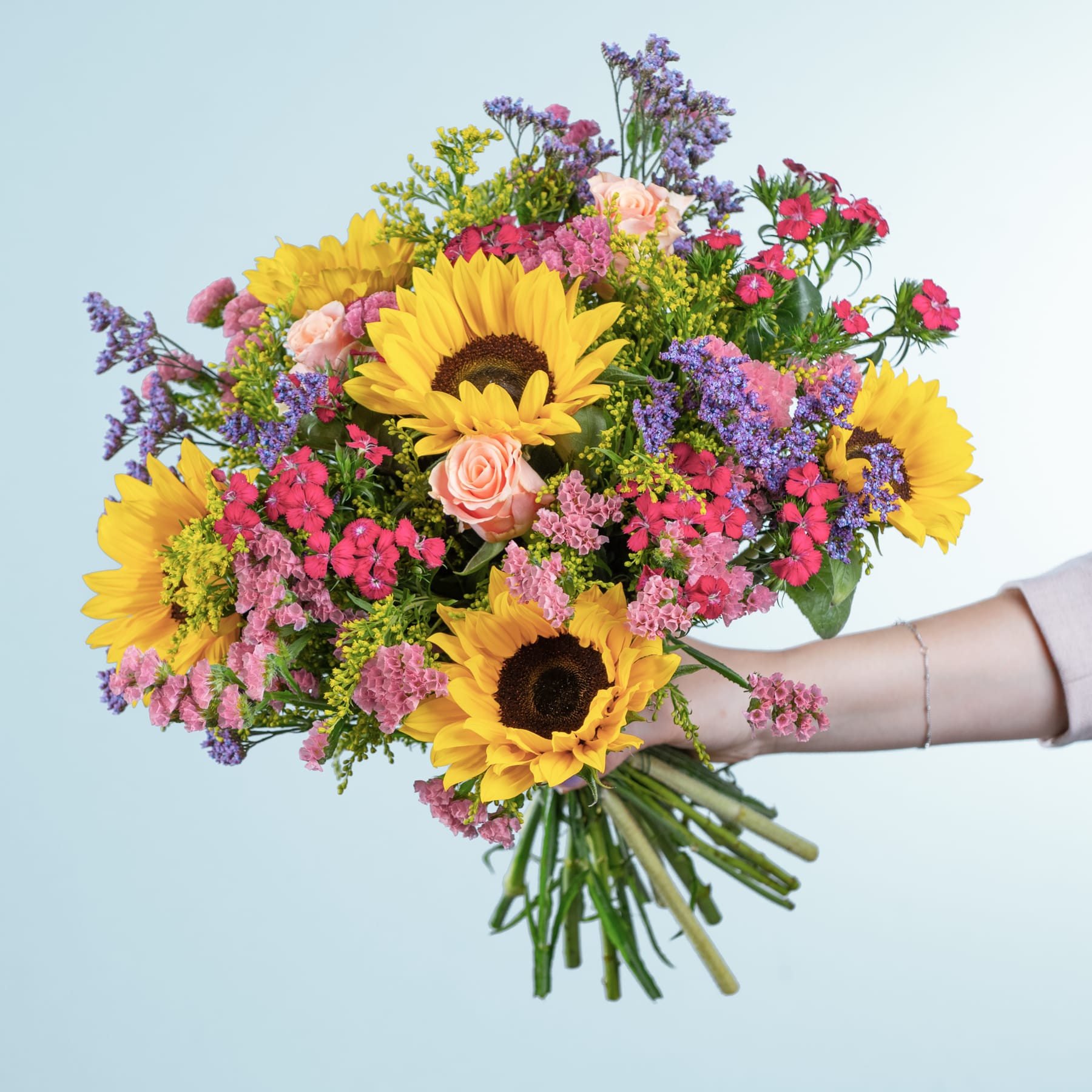 Ein leuchtender Strauß aus Sonnenblumen, Rosen und anderen bunten Blumen, den eine Person vor einem hellblauen Hintergrund in den Händen hält.