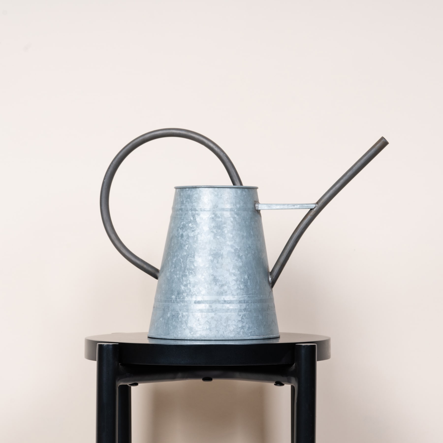 Eine Gießkanne aus verzinktem Metall steht auf einem schwarzen Metallständer vor einem schlichten beigen Hintergrund.