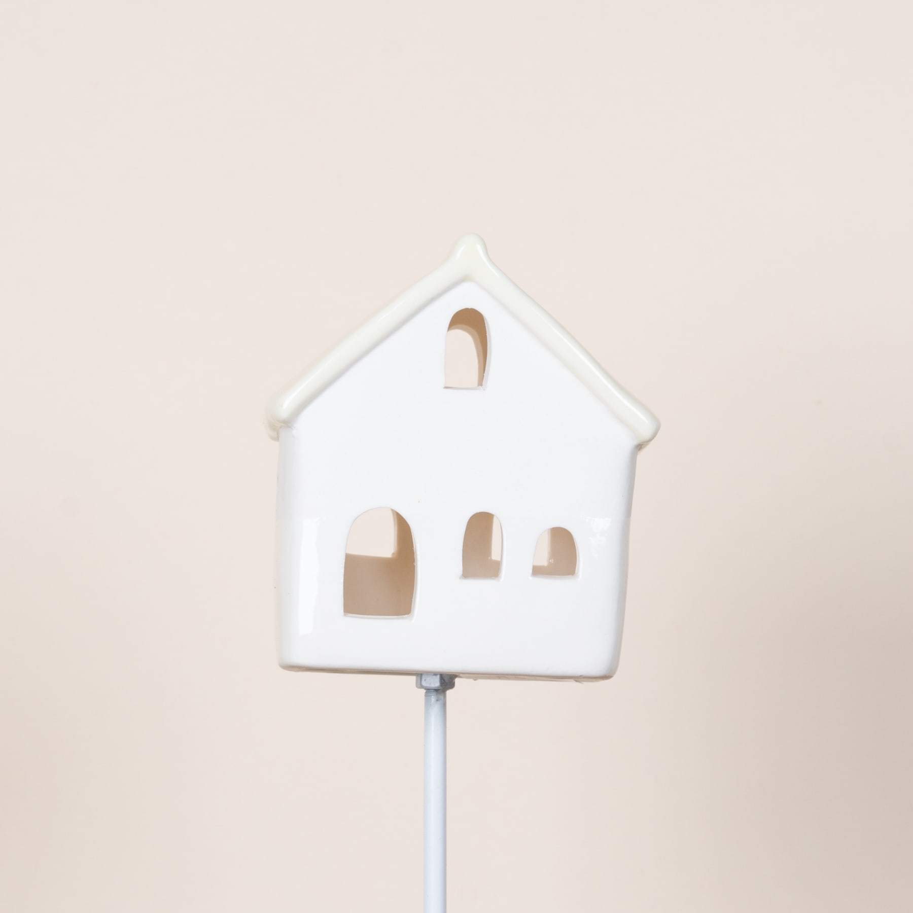 Vor einem schlichten, beigen Hintergrund steht ein kleines, weißes Keramikornament in Form eines Hauses mit ausgeschnittenen Fenstern und einem spitzen Dach.