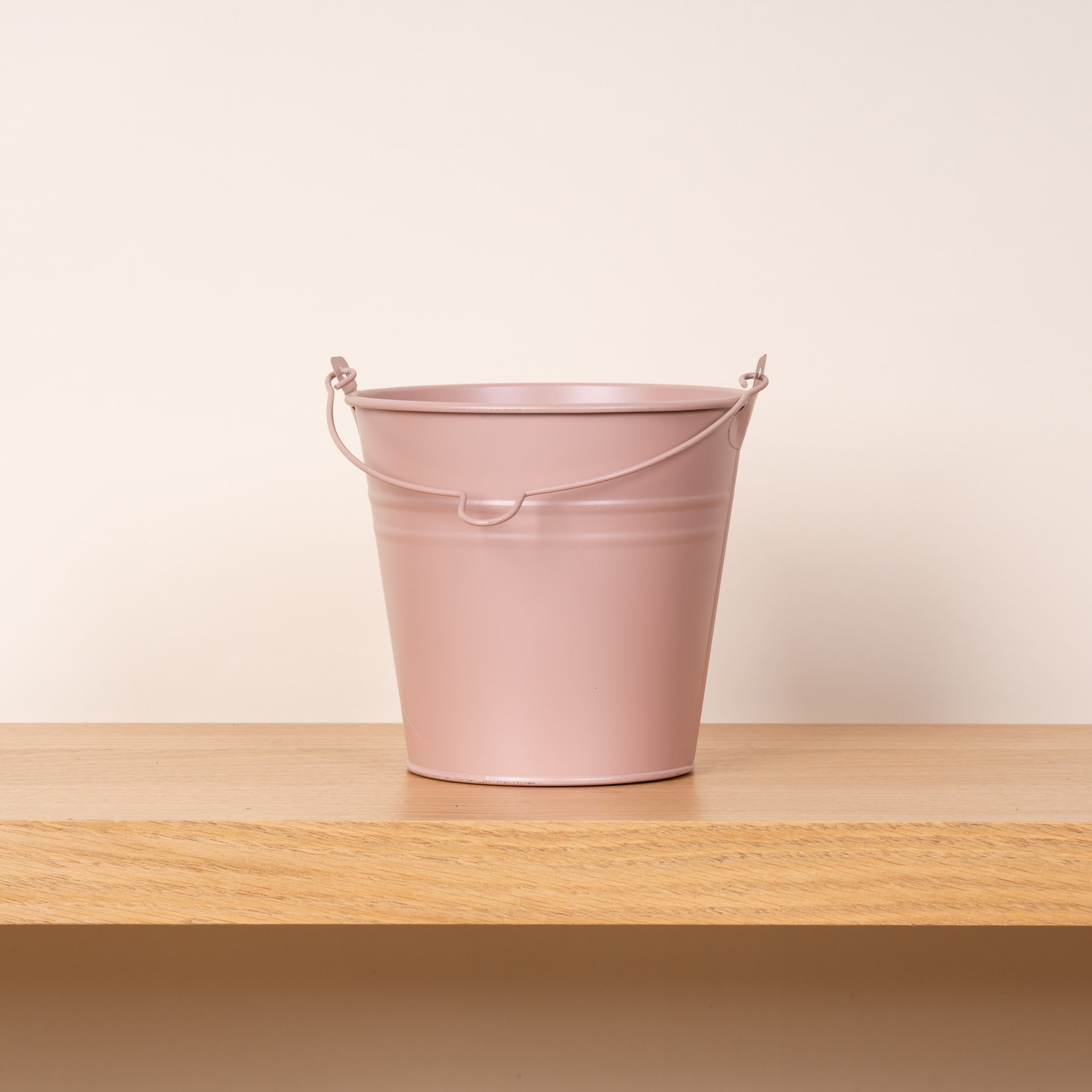 Ein rosa Metalleimer steht auf einem Holzregal vor einem schlichten, hellen Hintergrund.