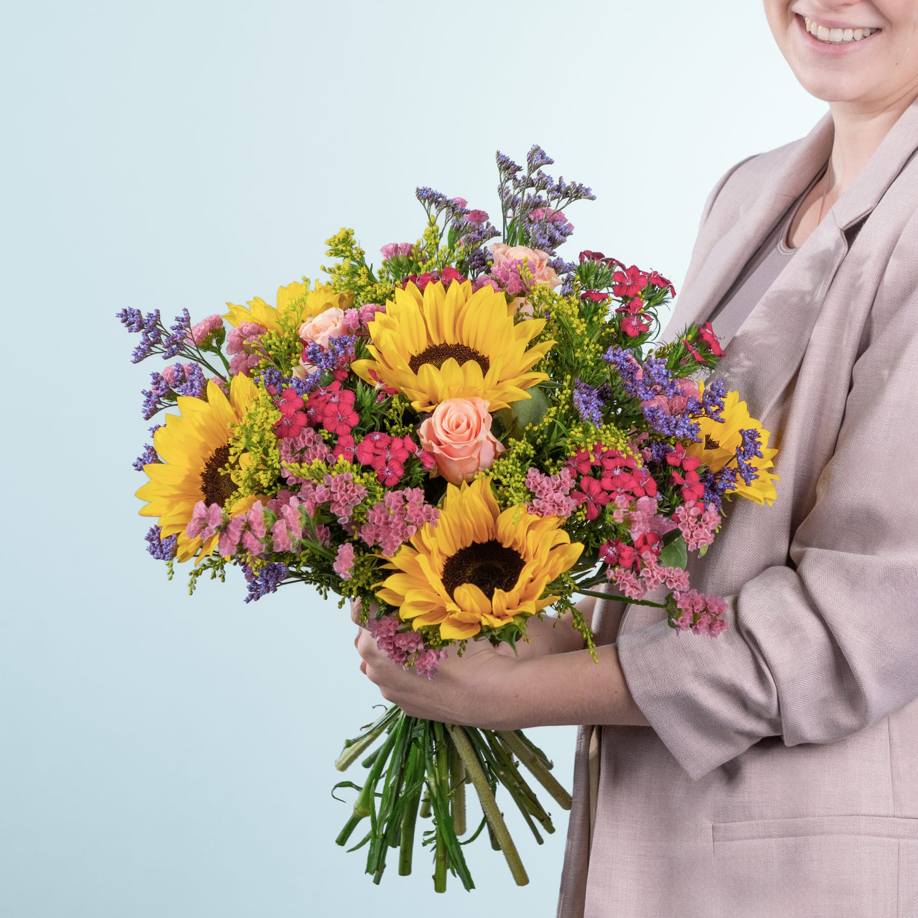 Ein leuchtender Strauß aus Sonnenblumen, Rosen und anderen bunten Blumen wird von einer Person gehalten, die einen beigen Mantel vor einem schlichten hellblauen Hintergrund trägt.