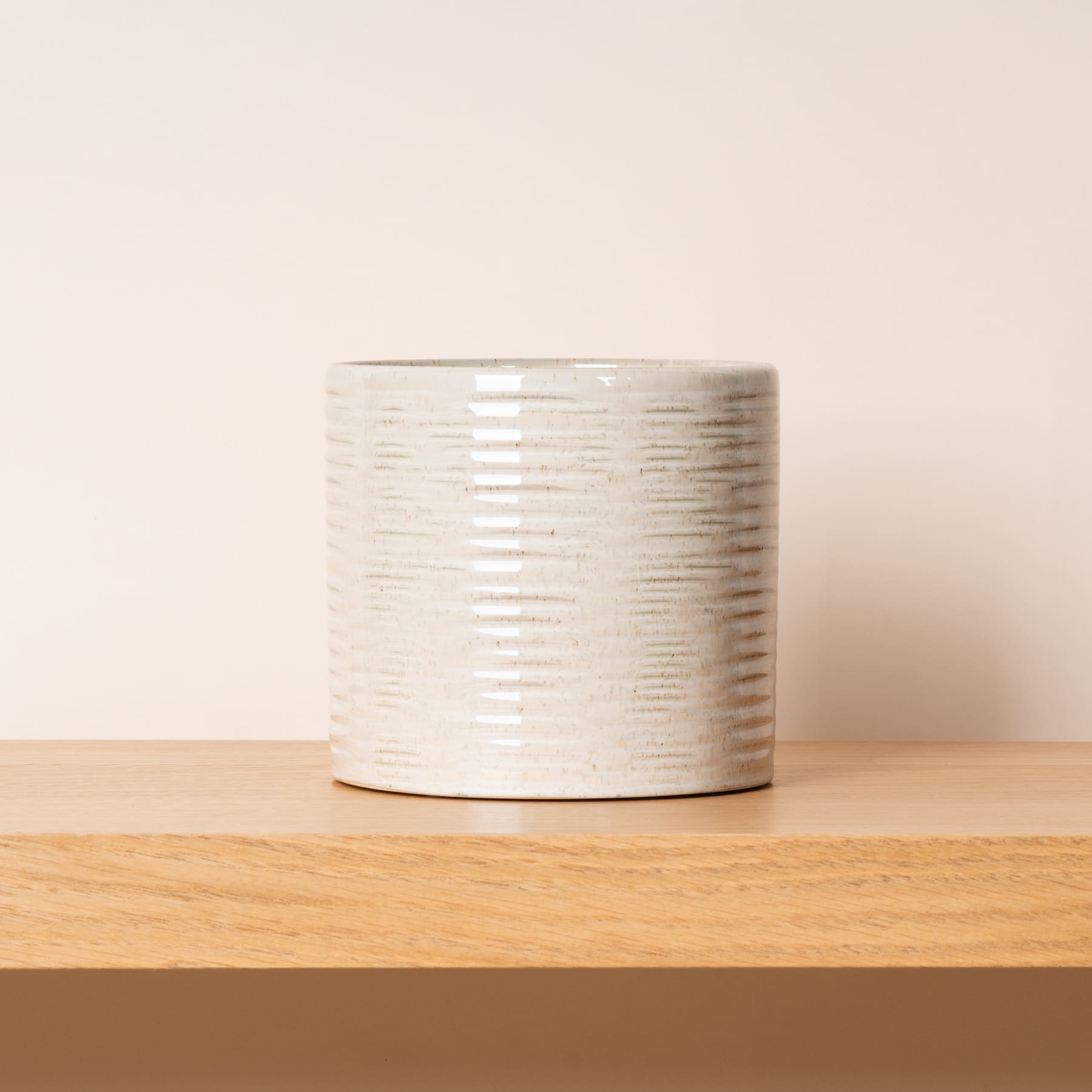 Ein weißer Blumentopf oder eine Vase aus Keramik mit strukturiertem Streifenmuster steht auf einer Holzoberfläche vor einem schlichten, hellen Hintergrund.