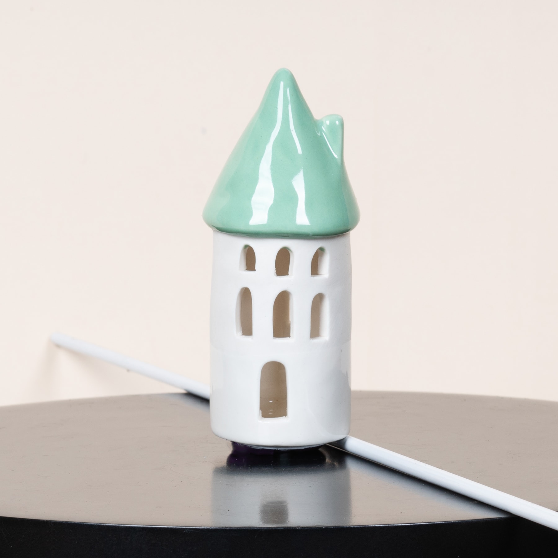 Ein kleines, weißes Keramikobjekt in Hausform mit mintgrünem Spitzdach steht auf einem dunklen Untergrund und reflektiert das Licht.