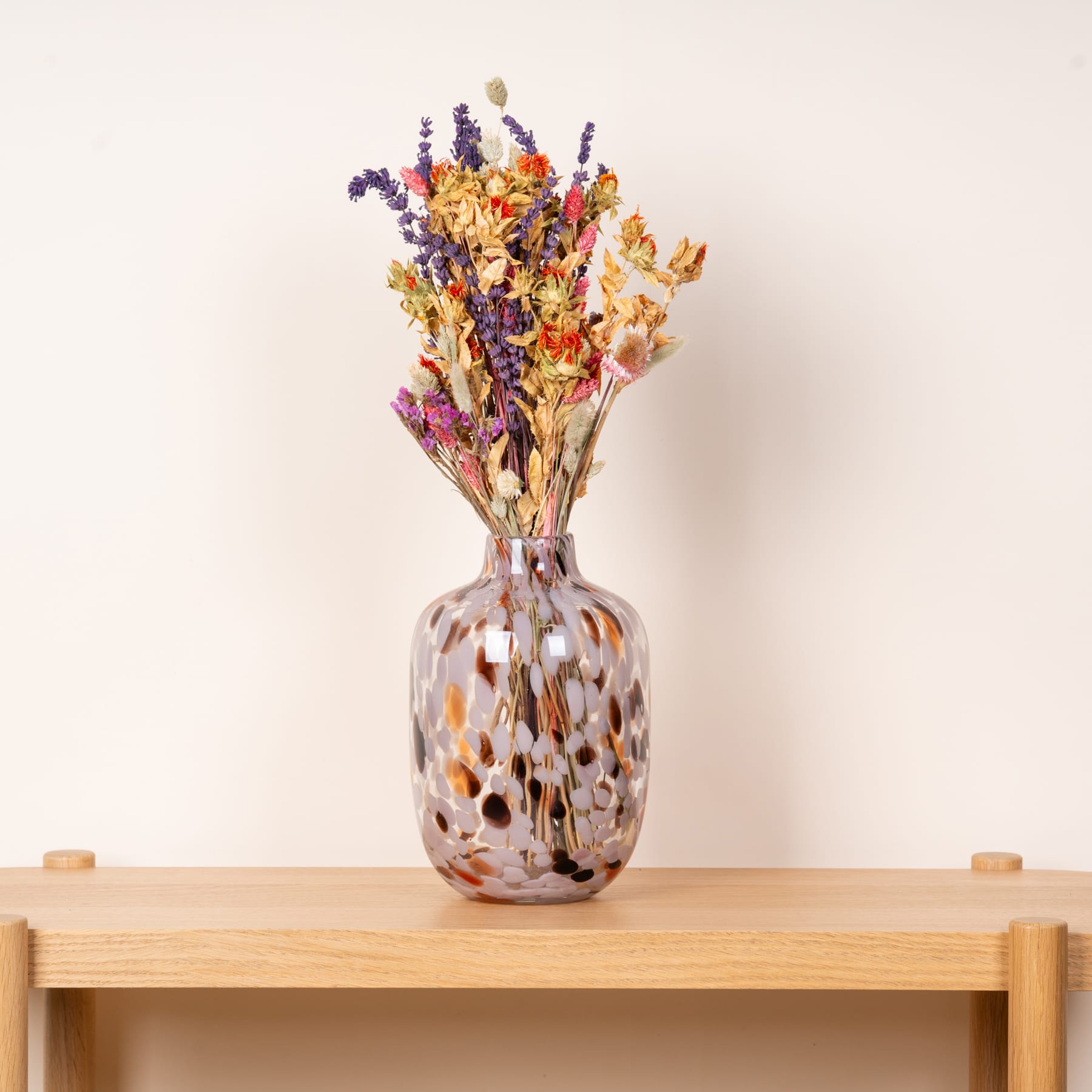 Eine Glasvase mit einem farbenfrohen Strauß getrockneter Blumen steht auf einem Holztisch vor einem schlichten beigen Hintergrund.