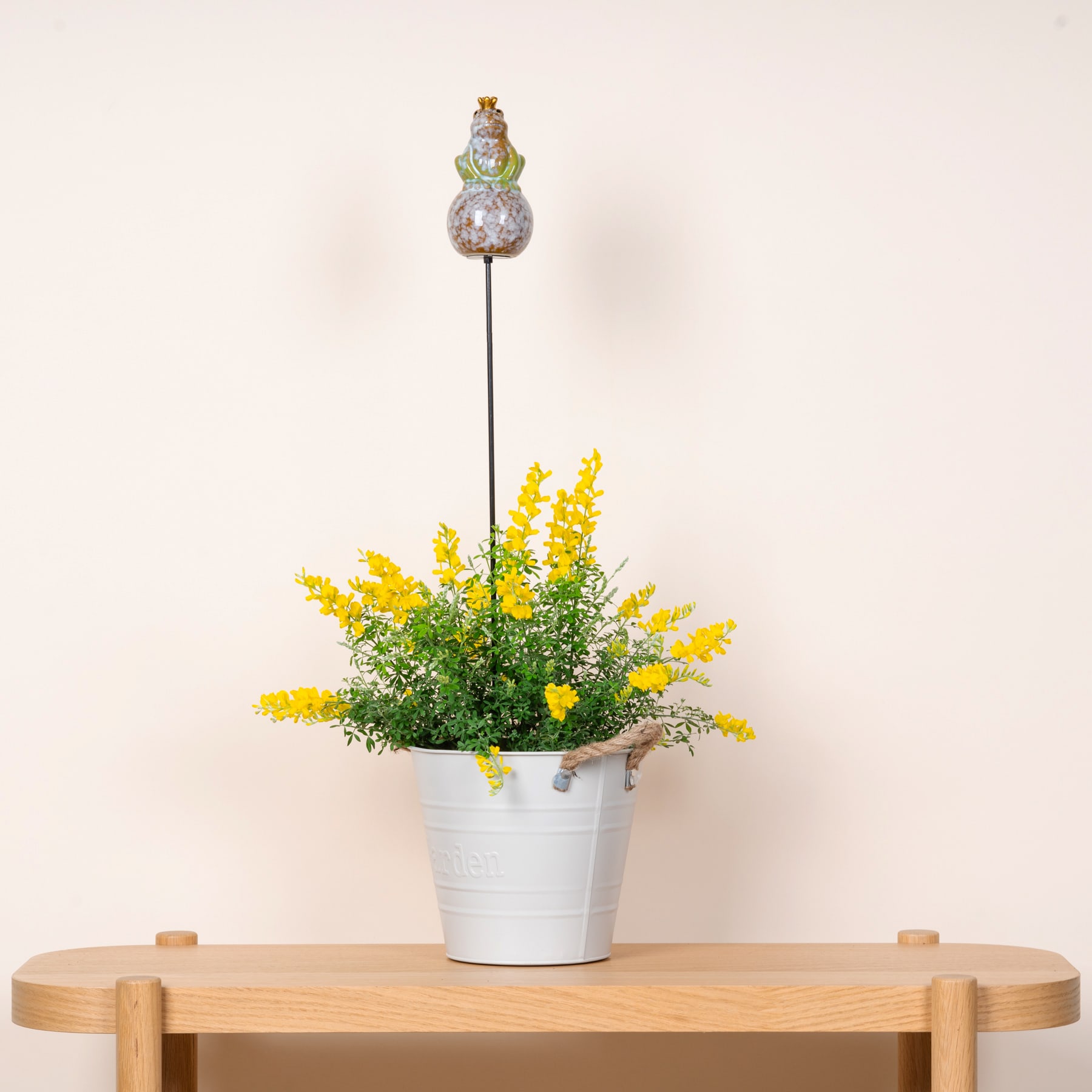 Ein weißer Metallbehälter mit leuchtend gelben Blumen steht auf einem Holztisch vor einem schlichten beigen Hintergrund.