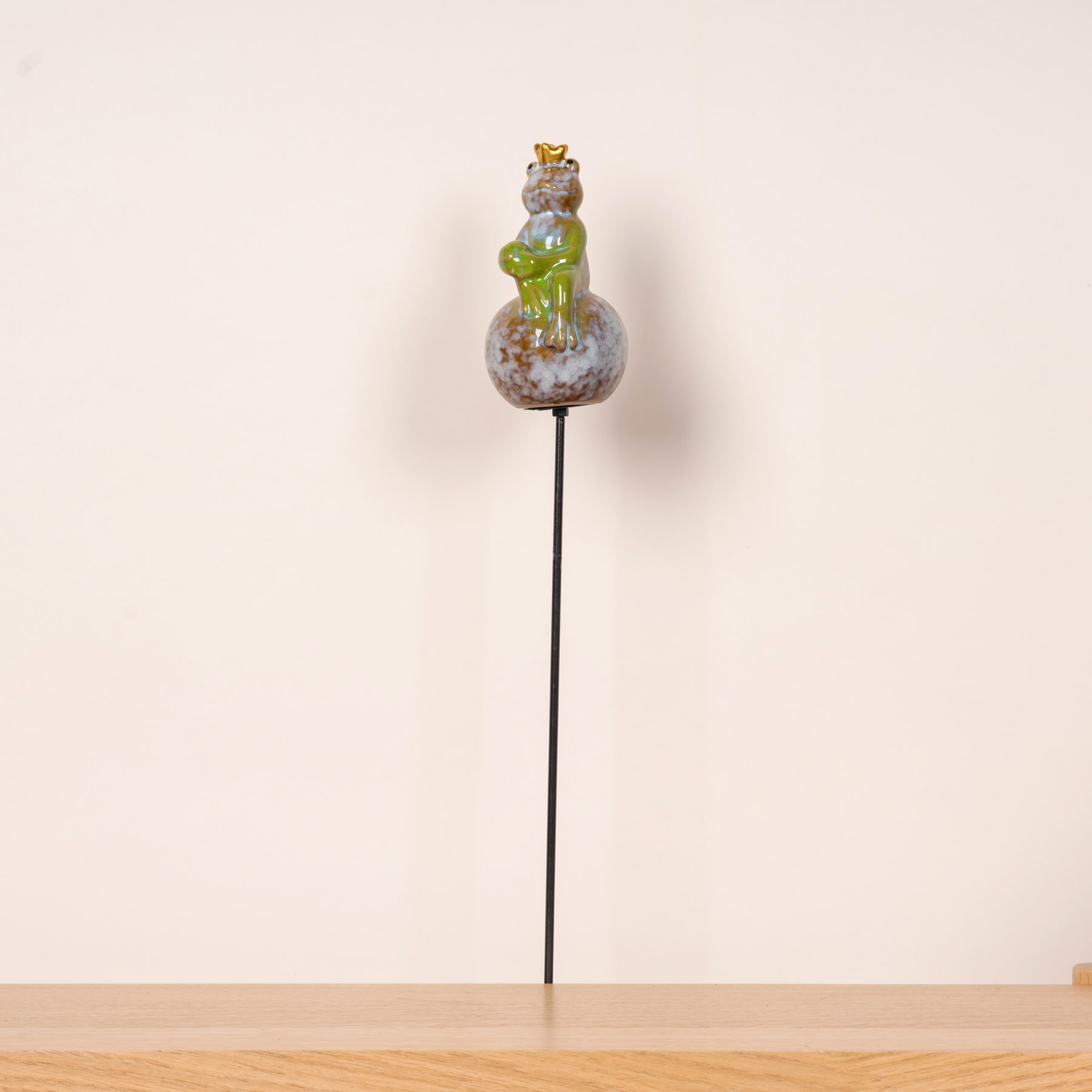 Auf einem schlichten, hellen Hintergrund sitzt auf einer Metallstange ein Glasornament in Froschform.