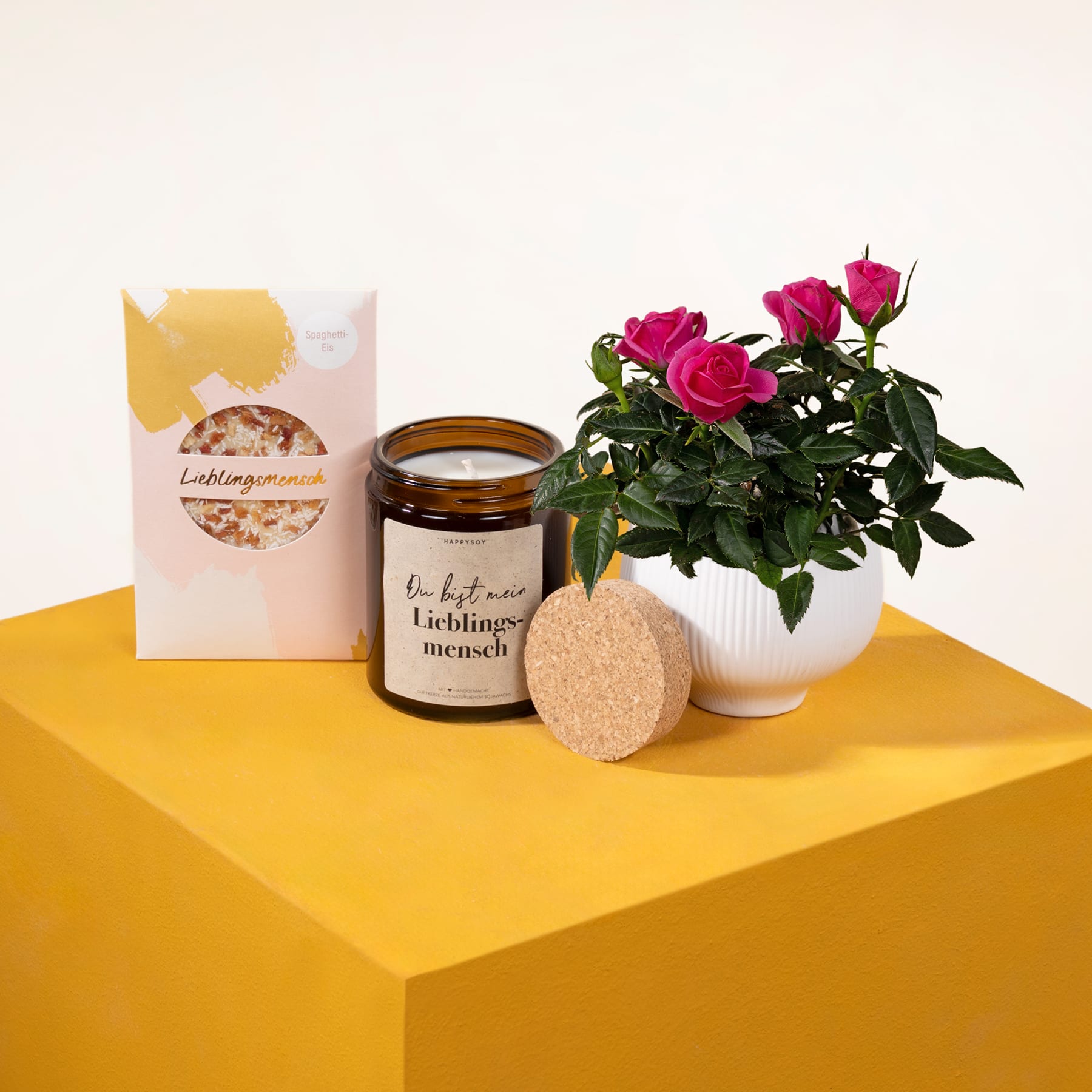 Auf einer gelben, würfelförmigen Plattform stehen ein Strauß leuchtend rosa Rosen, ein Glas Honig und eine Packung Müsli, alles vor einem schlichten weißen Hintergrund.