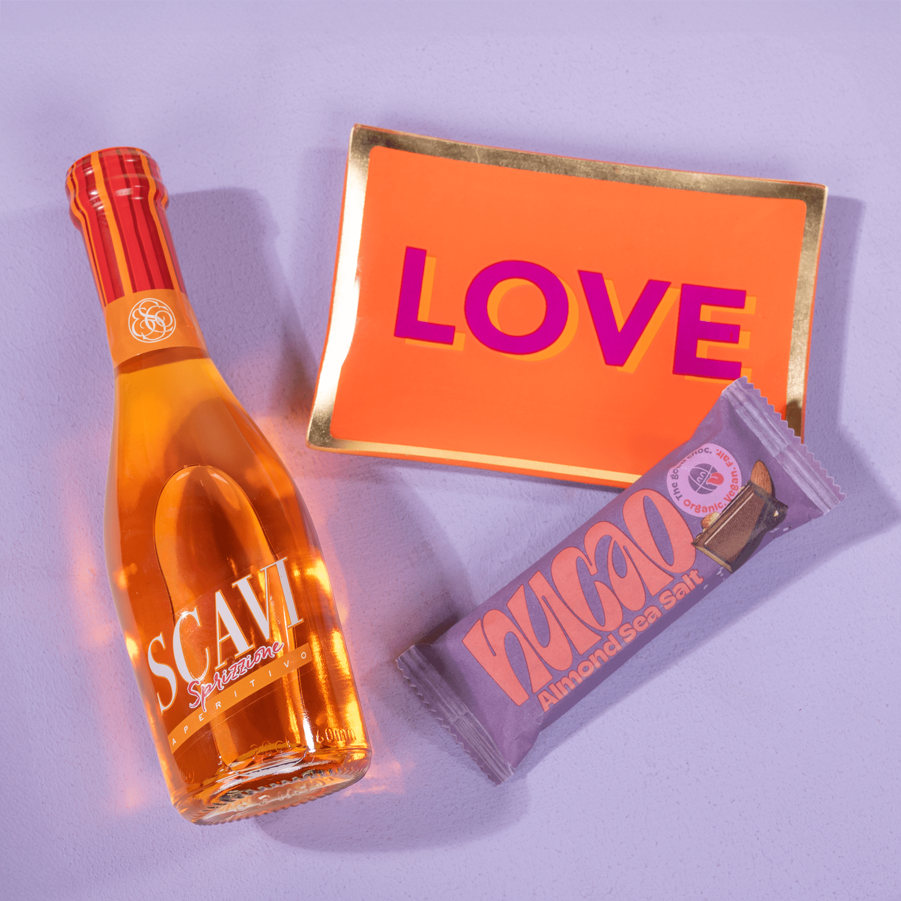 Das Bild zeigt eine orangefarbene Flasche Scam-Parfüm, ein rosa-orangefarbenes „LOVE“-Schild und einen lila-orangefarbenen Hagan-Schokoriegel vor einem lavendelfarbenen Hintergrund.