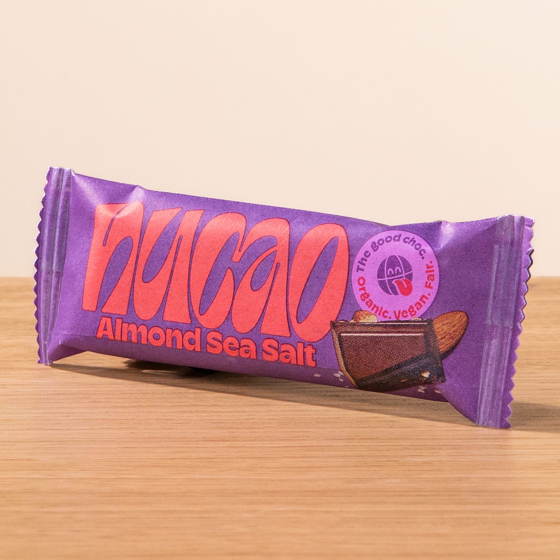 Eine lilafarbene Schokoriegelverpackung mit dem Wort „Nakao“ und der Geschmacksrichtung „Mandel-Meersalz“, die deutlich sichtbar auf einer Holzoberfläche abgebildet ist.