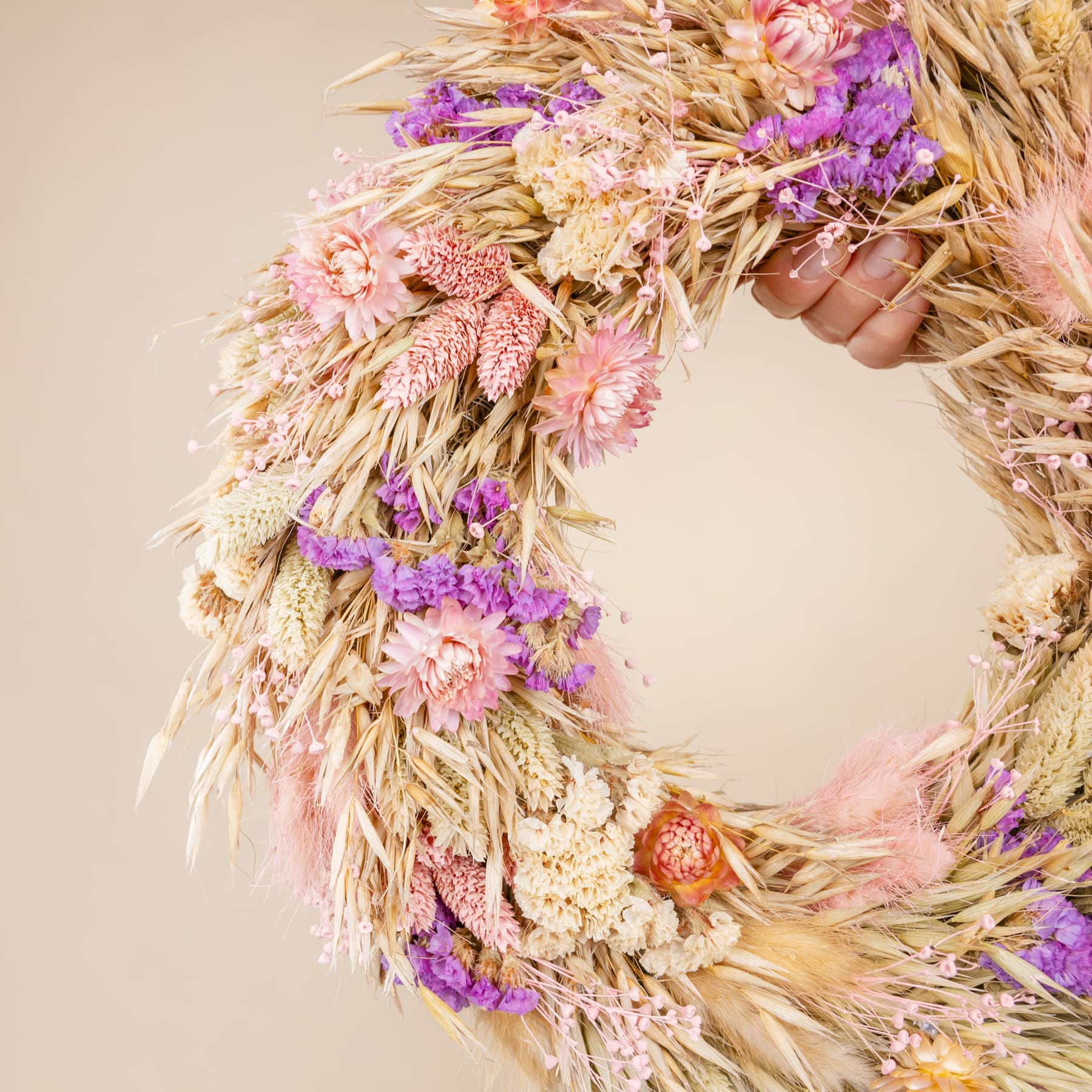 Ein farbenfroher Kranz aus getrockneten Blumen, Weizenhalmen und anderen natürlichen Elementen, der vor einem neutralen Hintergrund eine warme und rustikale Herbststimmung erzeugt.