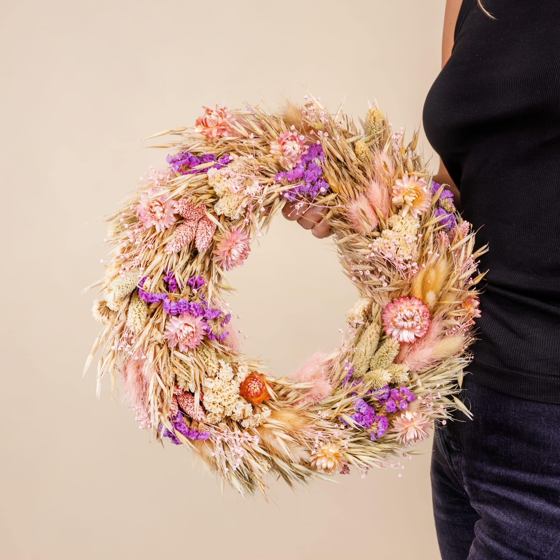 Ein leuchtender, runder Kranz aus getrockneten Blumen und Gräsern in Rosa-, Lila- und Gelbtönen, der vor einem neutralen Hintergrund vor den Körper einer Person gehalten wird.