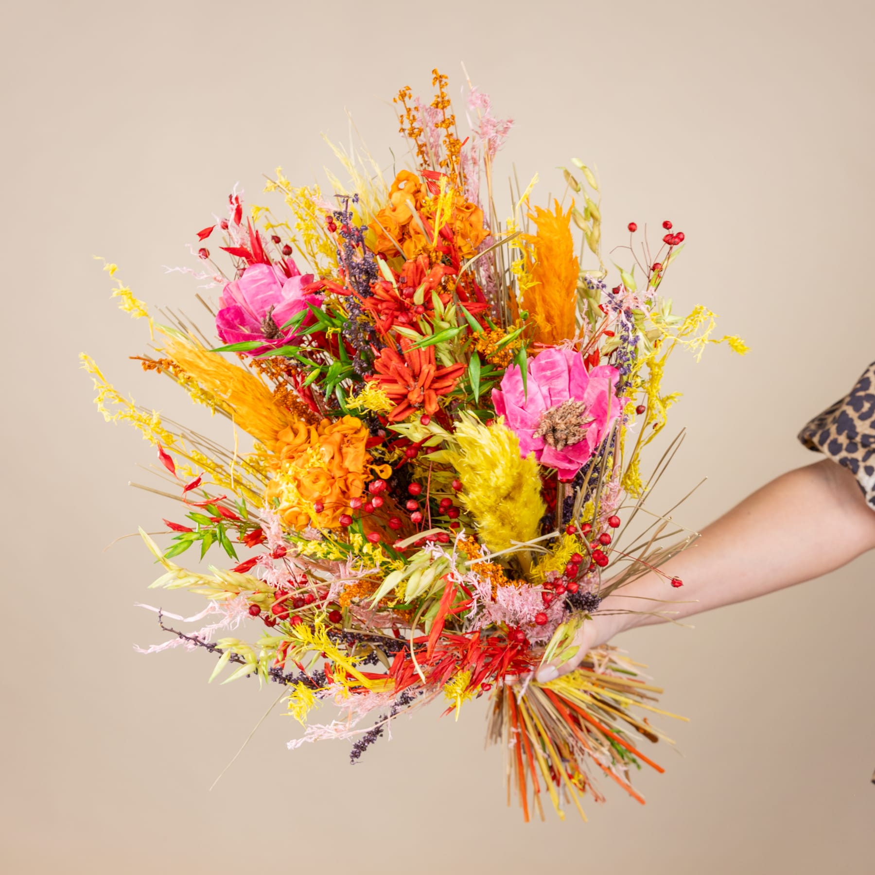 Ein lebendiger und farbenfroher Strauß verschiedener Blumen, darunter rosa, gelbe und orange Blüten, steht im Vordergrund vor einem neutralen beigen Hintergrund.
