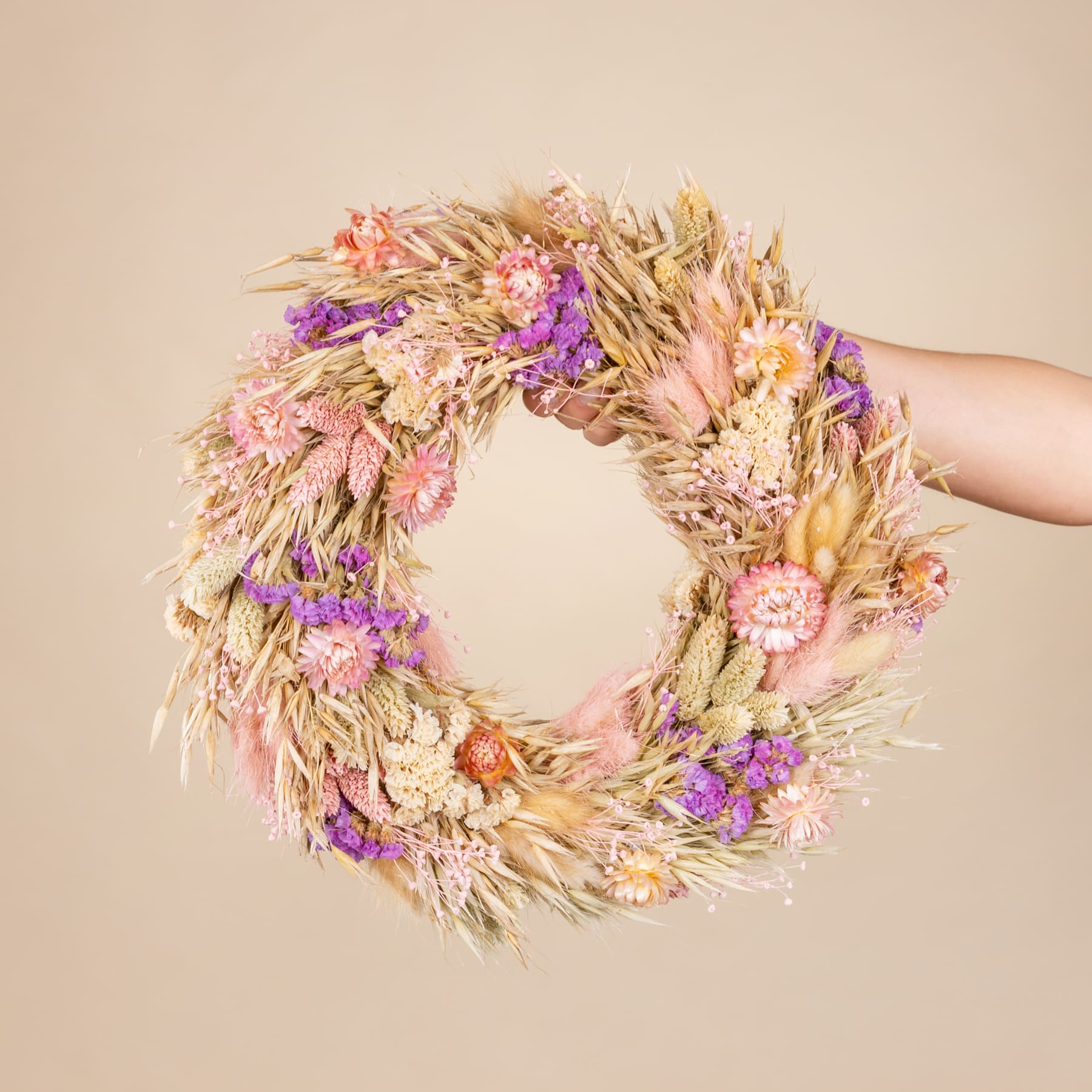 Ein wunderschöner Kranz aus getrockneten Blumen und Weizenhalmen, der vor einem neutralen beigen Hintergrund in den Händen einer Person gehalten wird.