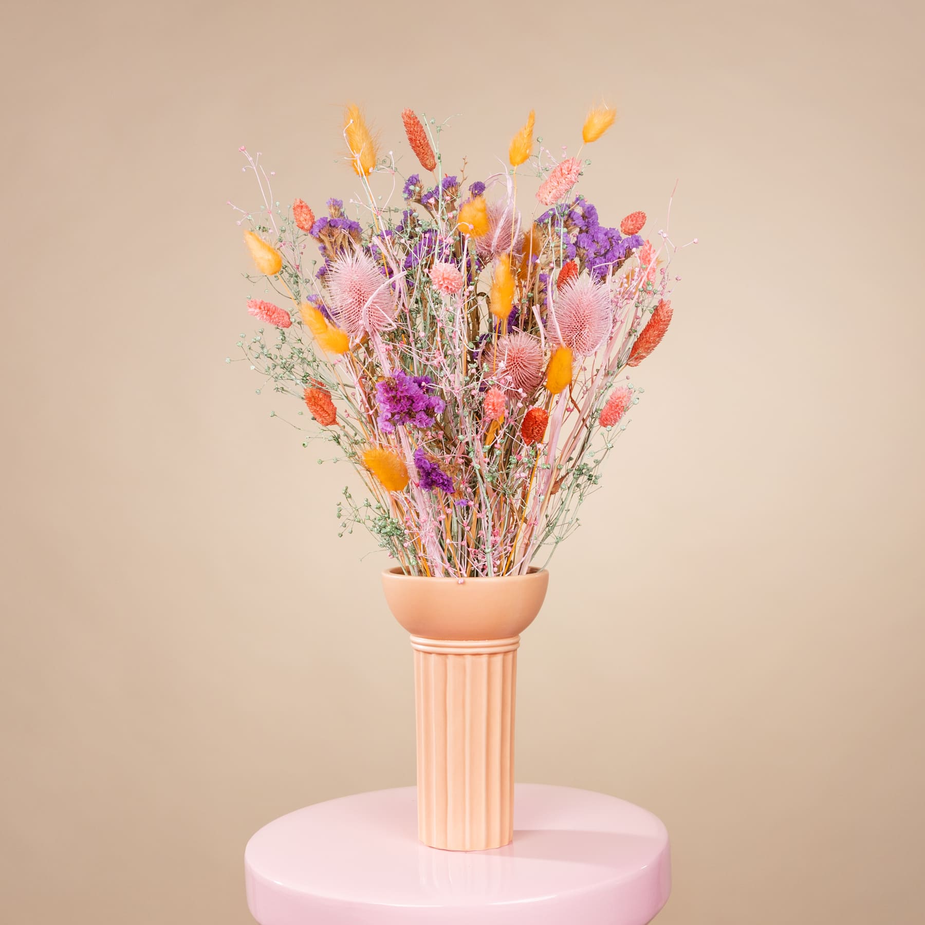 Ein lebendiger Strauß bunter Trockenblumen in einer Terrakottavase steht auf einem rosa Sockel vor einem neutralen beigen Hintergrund.