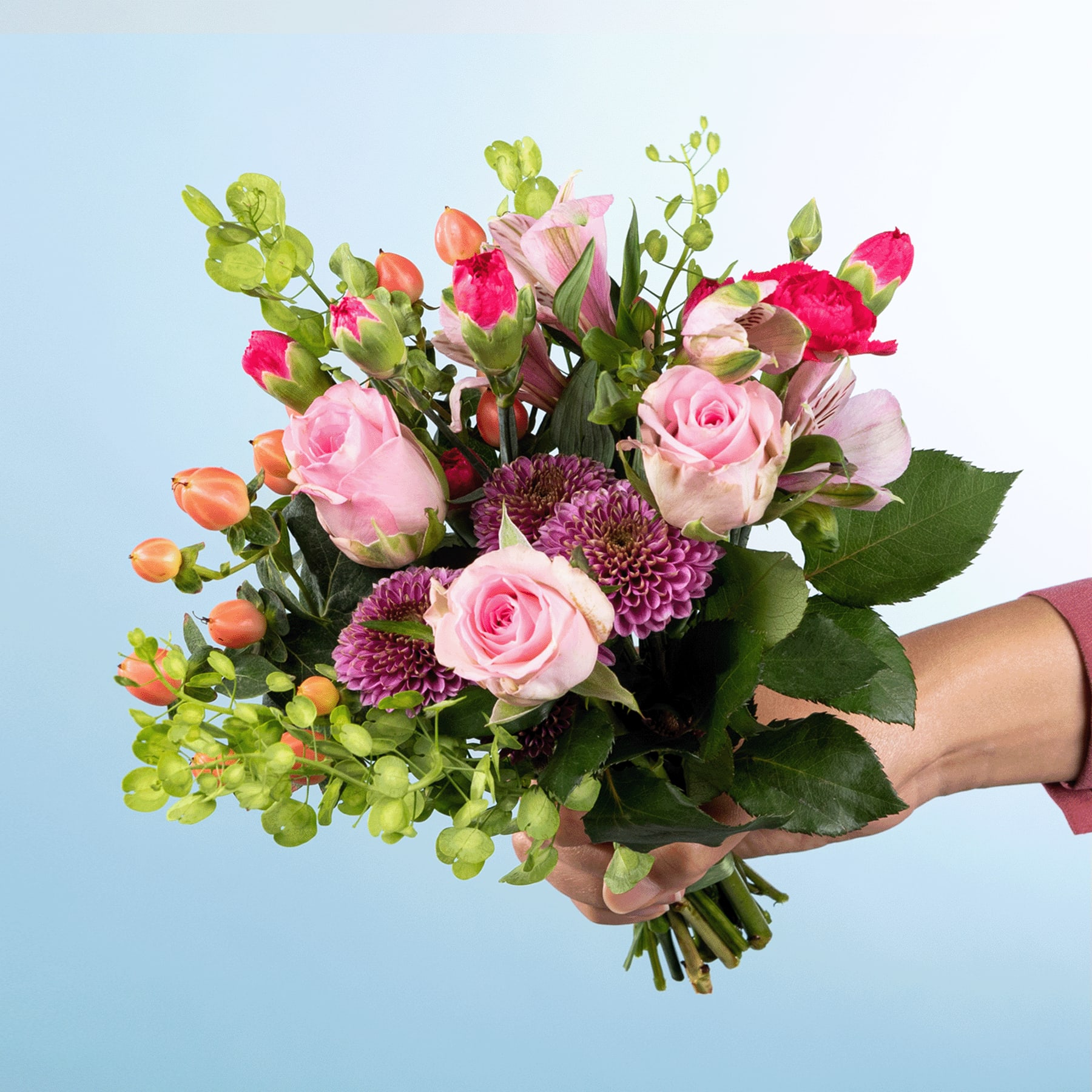 Ein leuchtender Strauß aus rosa und roten Rosen, Chrysanthemen und anderen Grünpflanzen, den eine Person vor einem hellblauen Hintergrund in den Händen hält.