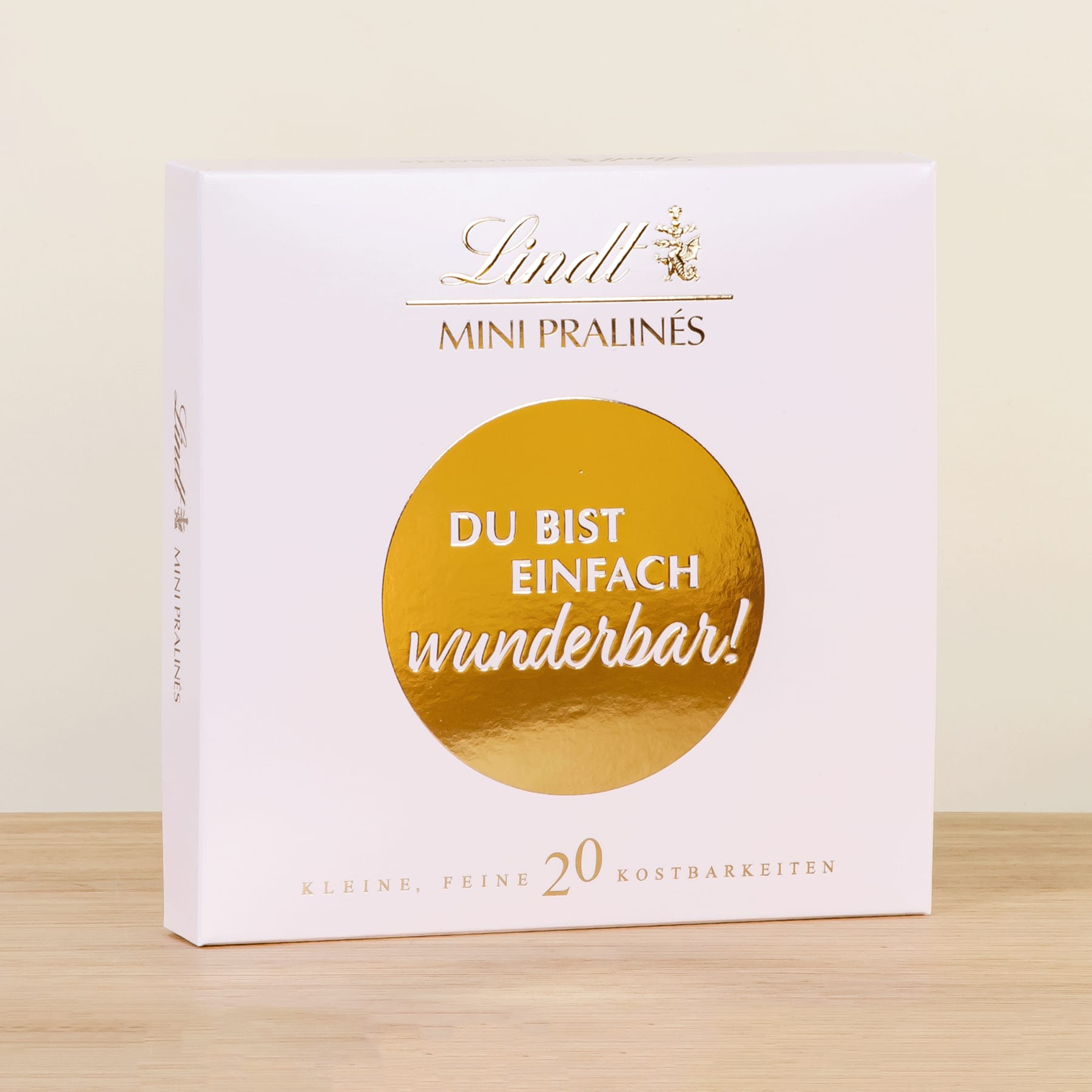 Eine weiße Schachtel mit dem Lindt-Logo und dem Text „Du bist einfach wunderbar!“ in einem goldenen Kreis auf der Vorderseite, stehend auf einer Holzoberfläche.