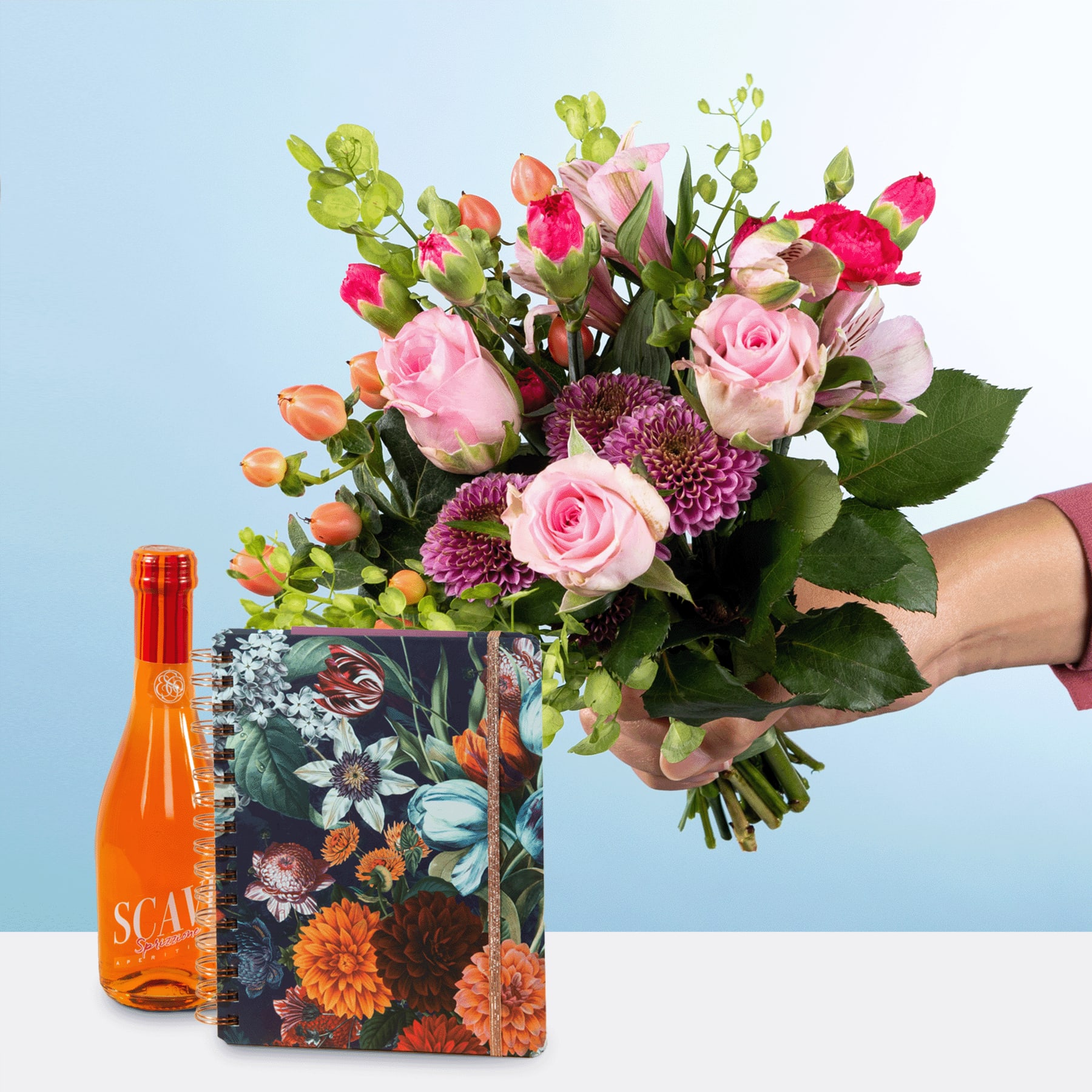 Ein leuchtender Strauß aus rosa Rosen, lila Chrysanthemen und anderen bunten Blumen wird vor einem hellblauen Hintergrund vor einer Flasche mit orangefarbener Flüssigkeit gehalten.