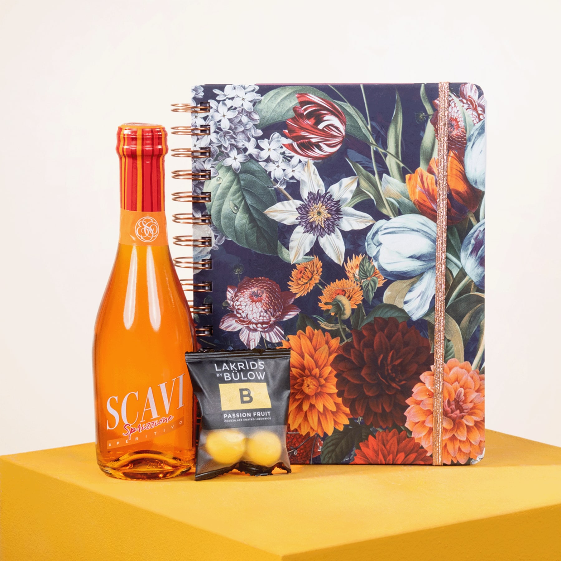 Ein farbenfrohes Notizbuch mit Blumenmuster, eine orangefarbene Flasche und eine Zitrone stehen auf einer gelben Oberfläche und ergeben ein optisch auffälliges und farbenfrohes Arrangement.
