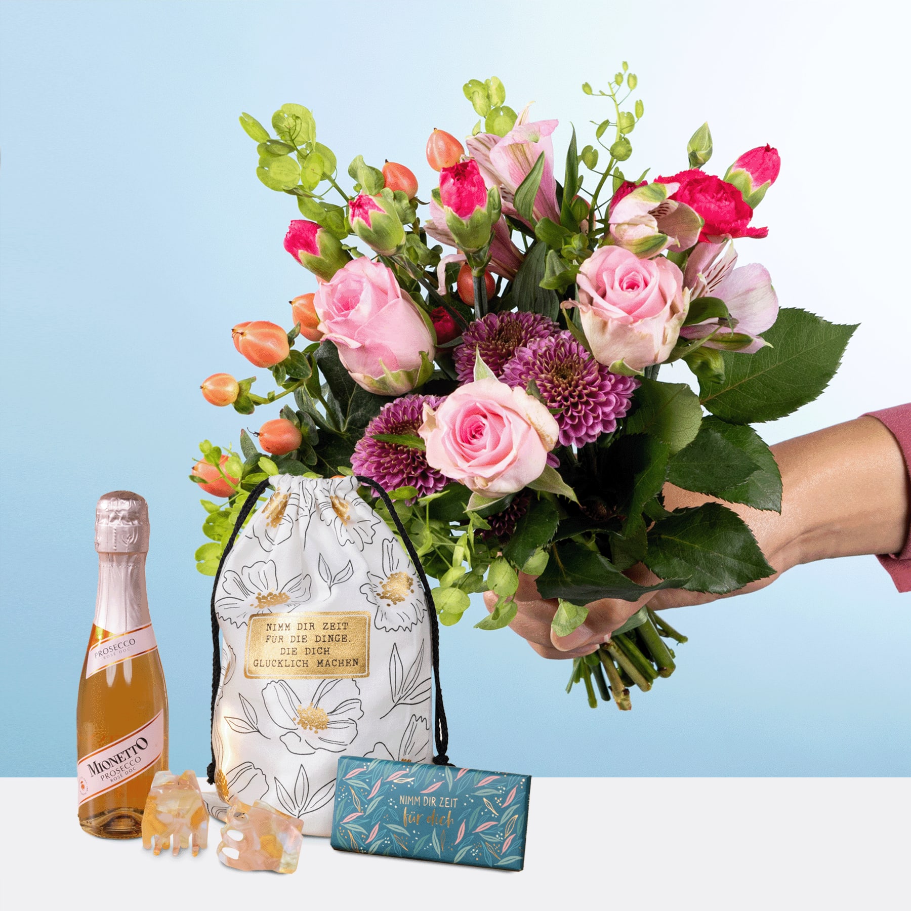 Ein lebendiger Strauß aus rosa Rosen, lila Chrysanthemen und anderen bunten Blumen, gehalten von einer Hand, begleitet von einer Flasche Sekt und einer Geschenkbox auf hellblauem Hintergrund.