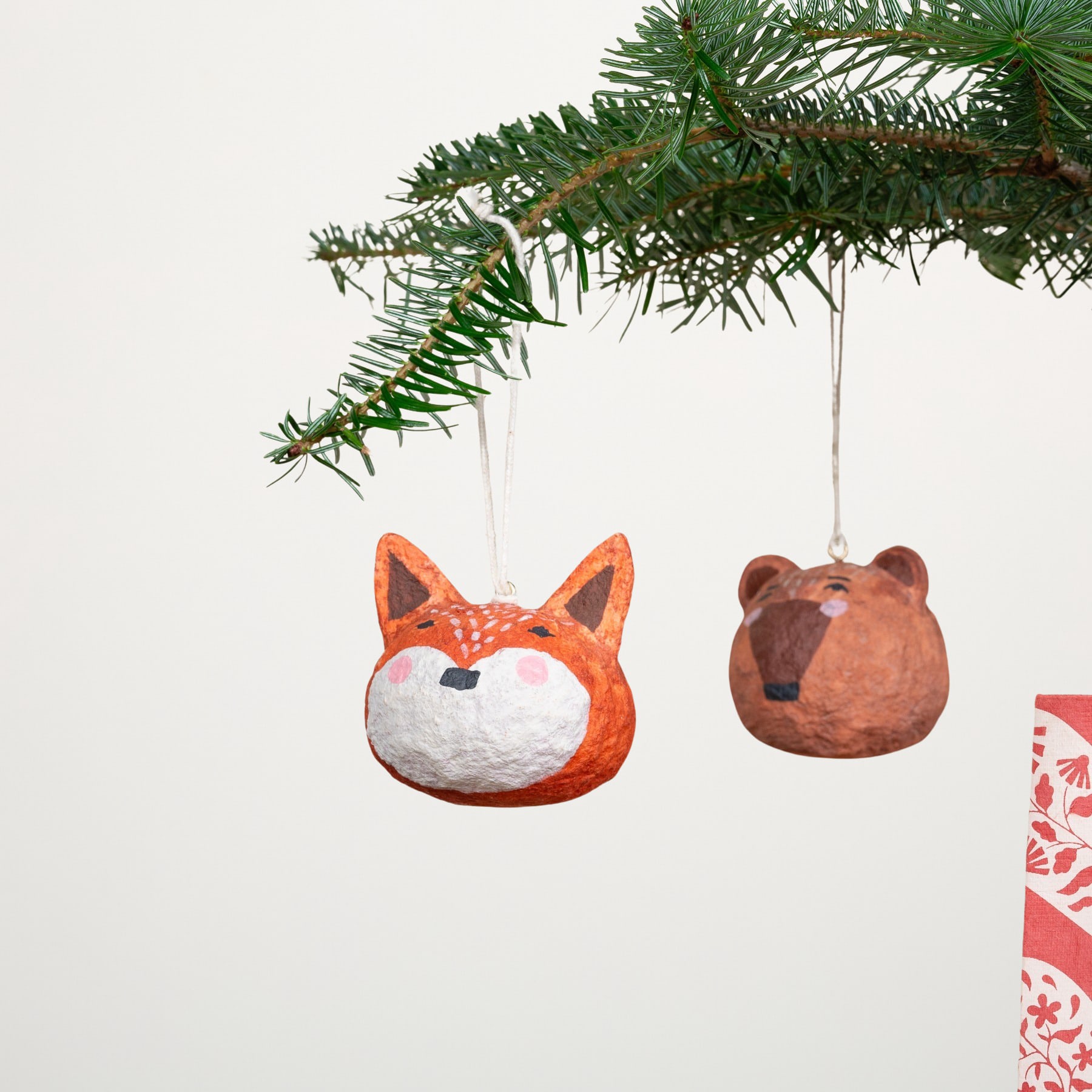 Das Bild zeigt zwei Ornamente, die an einem Weihnachtsbaumzweig hängen – eines in Form eines Fuchses und das andere in Form eines Bären.