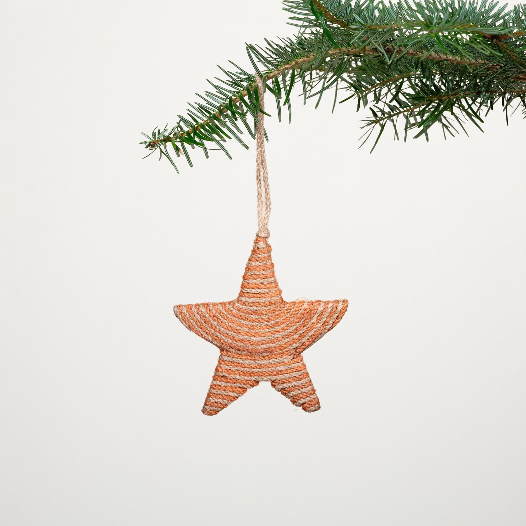 An einem Kiefernzweig hängt ein geflochtenes, sternförmiges Ornament, dessen warmer Orangeton einen Kontrast zu den üppigen grünen Nadeln bildet, die es umgeben.