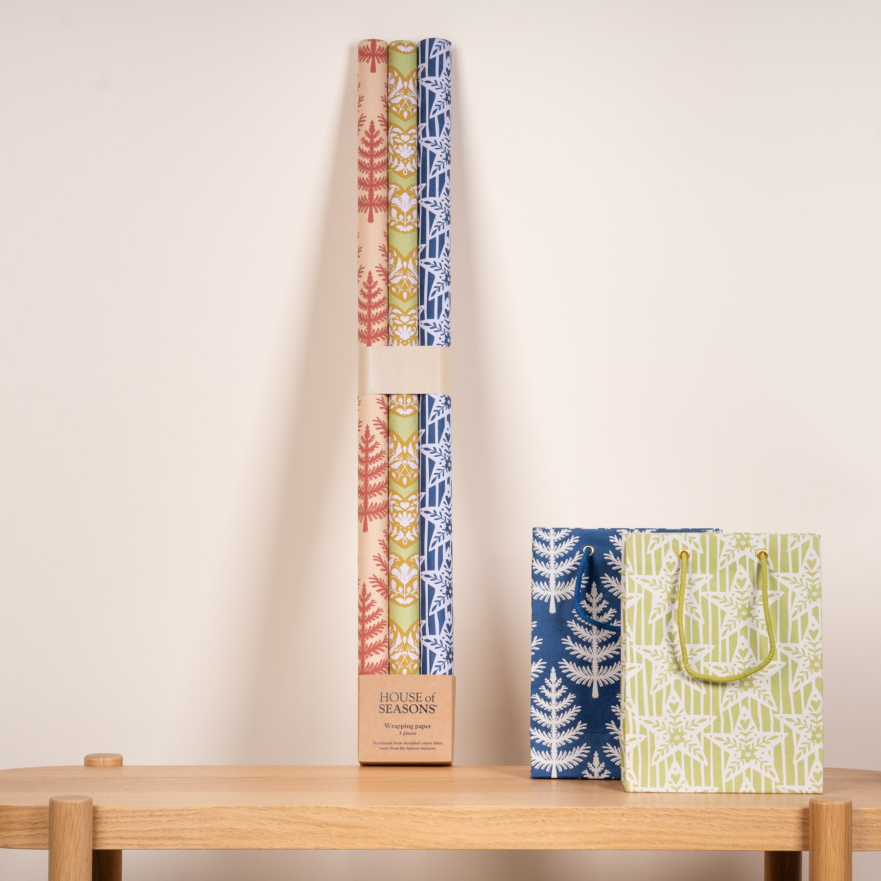 Das Bild zeigt mehrere Rollen gemustertes Geschenkpapier und eine Geschenktüte auf einer Holzoberfläche vor einem schlichten Hintergrund.