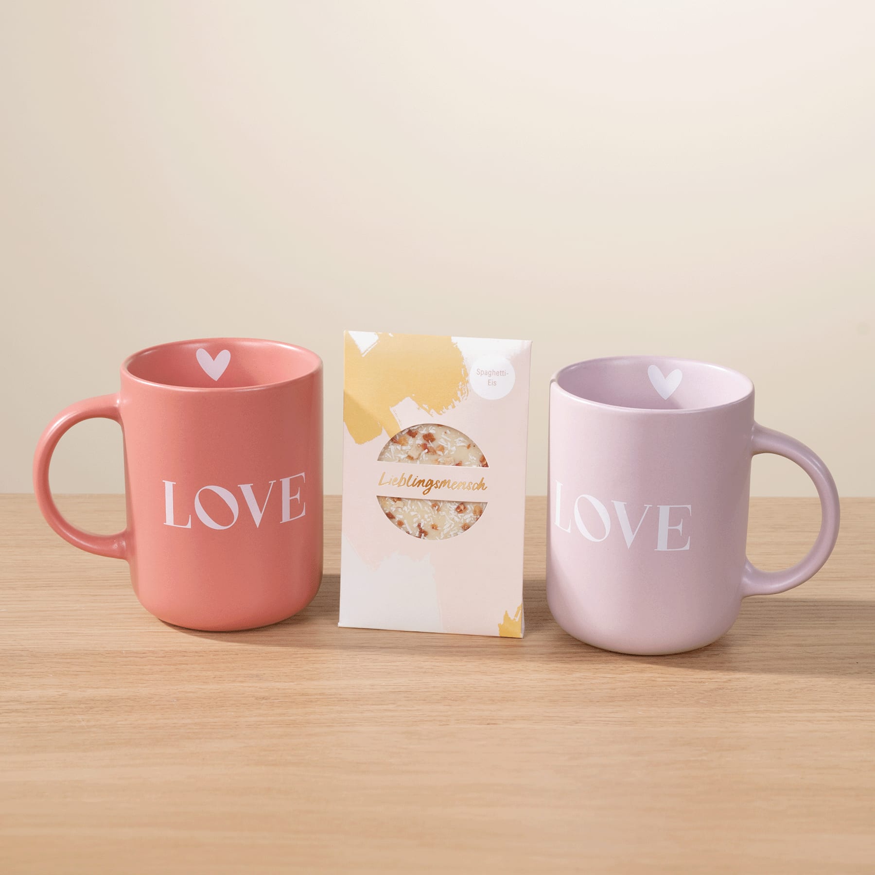 Zwei pastellfarbene Tassen mit dem Aufdruck „LOVE“ und eine kleine weiße Geschenkbox stehen auf einer Holzfläche vor einem hellen Hintergrund.