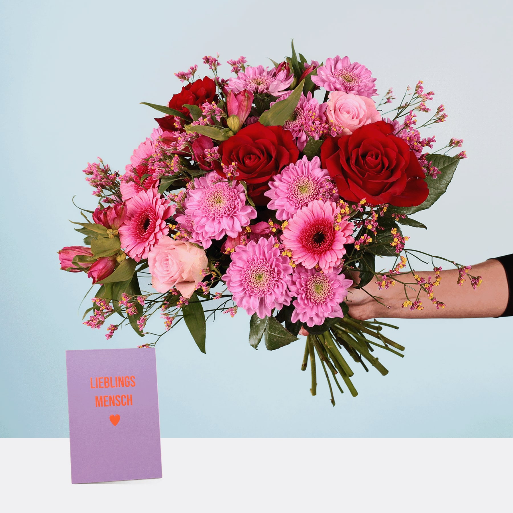 Ein leuchtender Strauß roter Rosen, rosa Gänseblümchen und anderer bunter Blumen in den Händen einer Person, mit einer kleinen Karte mit der Aufschrift „Geluks wens“ (niederländisch für „Herzlichen Glückwunsch“) im Vordergrund vor einem schlichten blauen Hintergrund.