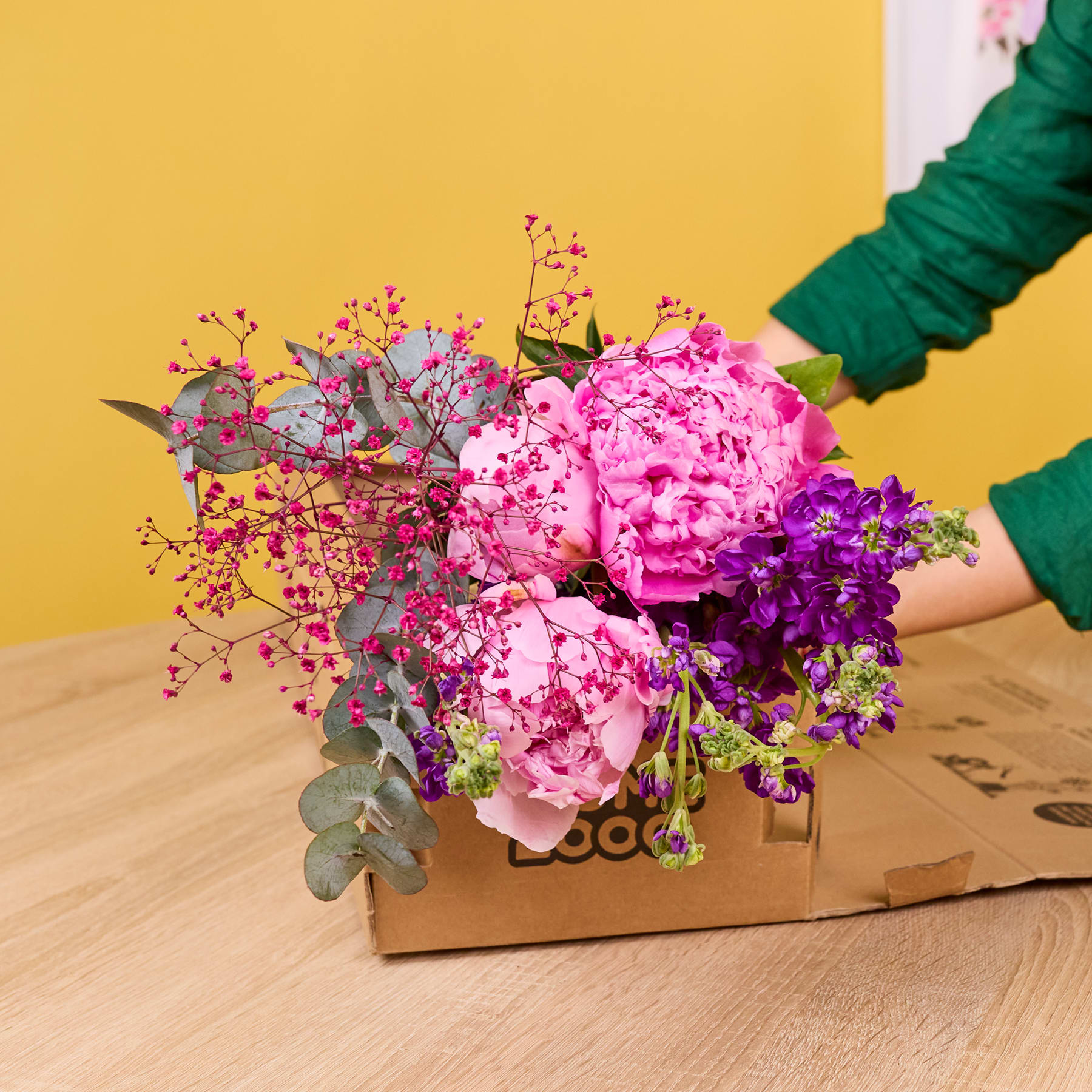 Ein farbenfroher Strauß aus rosa Pfingstrosen, violetten Schwertlilien und anderen bunten Blumen ist in einer Holzkiste arrangiert und steht auf einer Holzoberfläche vor einem leuchtend gelben Hintergrund.