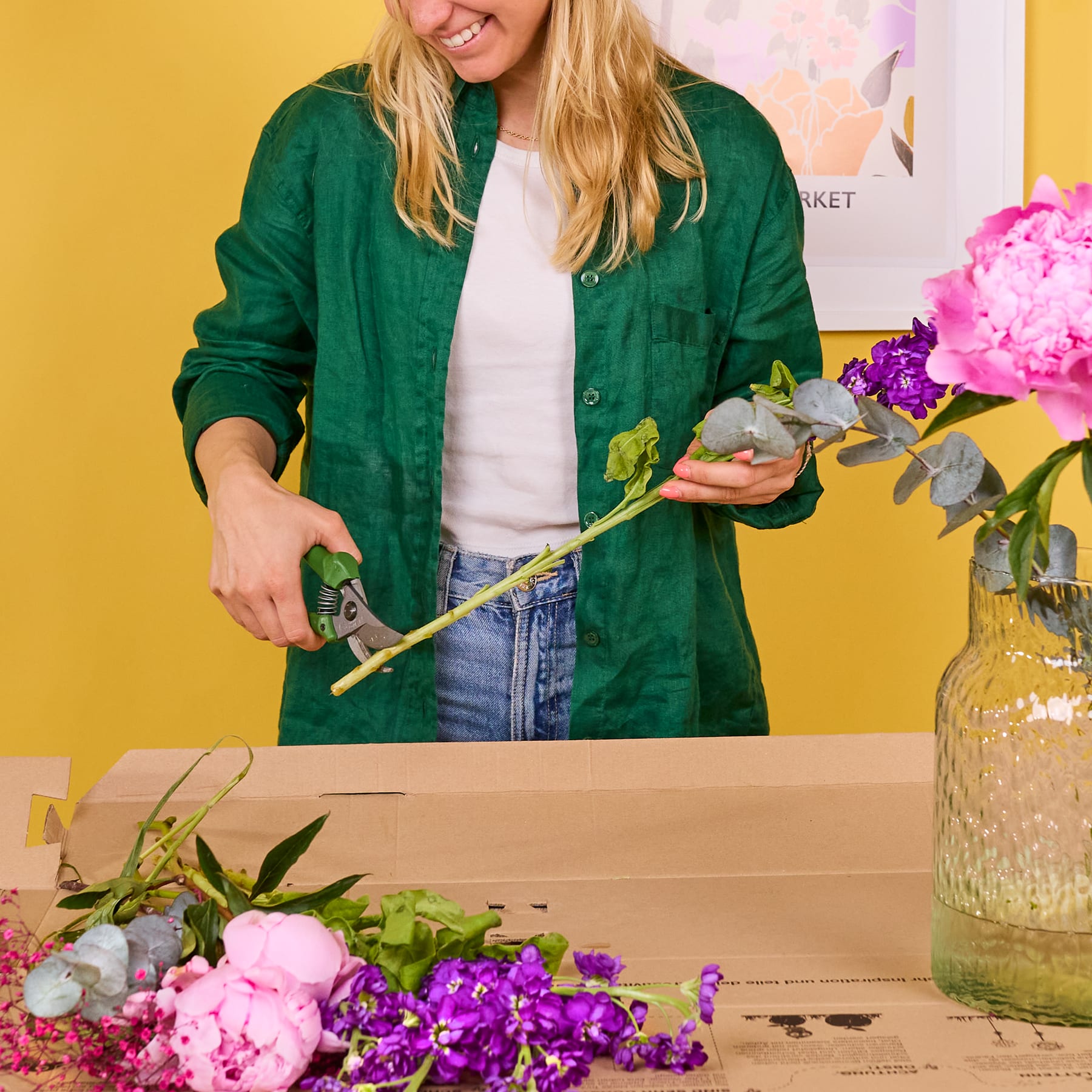 Eine Frau in einer grünen Jacke arrangiert Blumen in einer Vase auf einem Tisch mit einem farbenfrohen Blumenarrangement und gelbem Hintergrund.