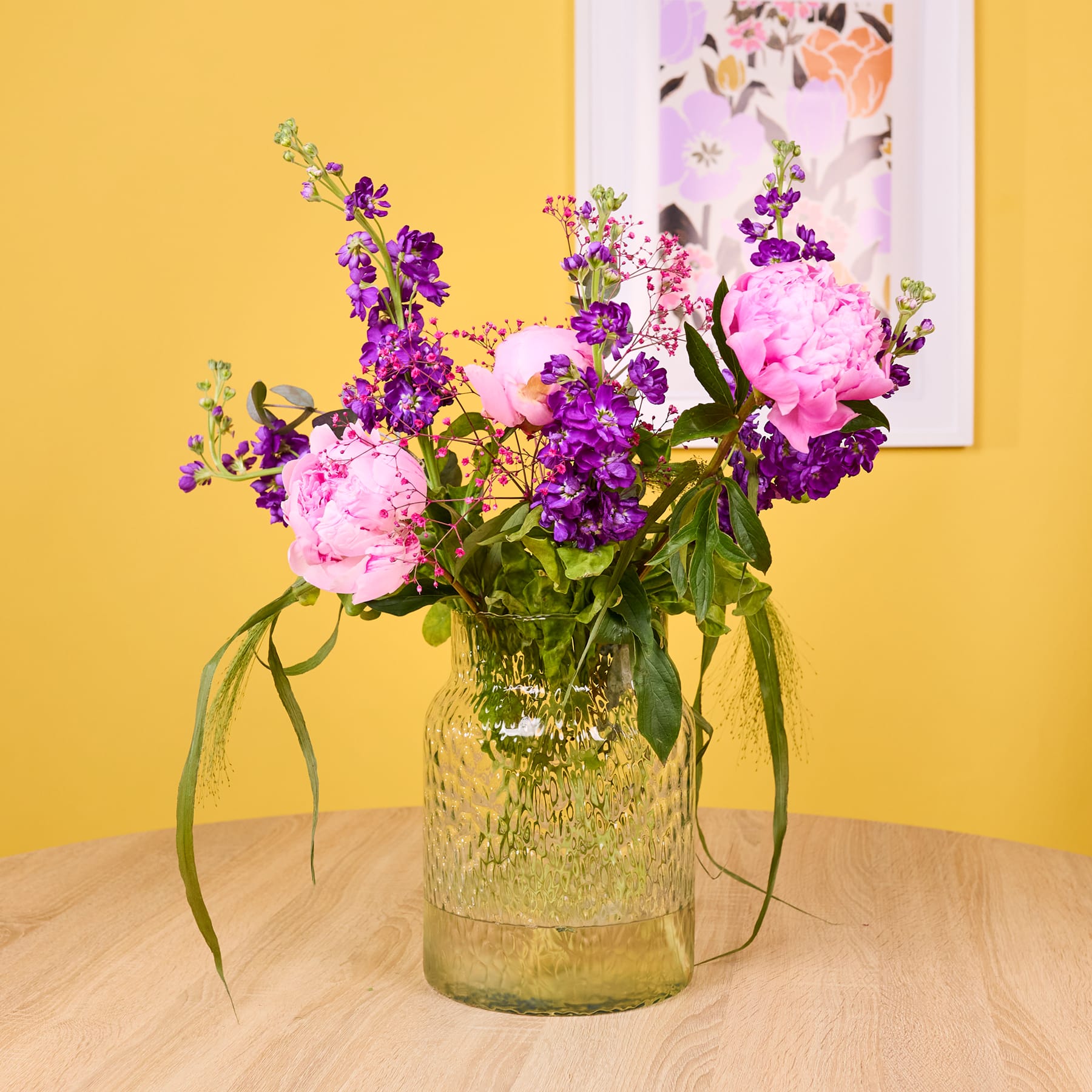 Ein farbenfroher Strauß aus rosa Pfingstrosen, violetten Schwertlilien und anderen bunten Blumen steht in einer Glasvase auf einem Holztisch vor einem leuchtend gelben Hintergrund.