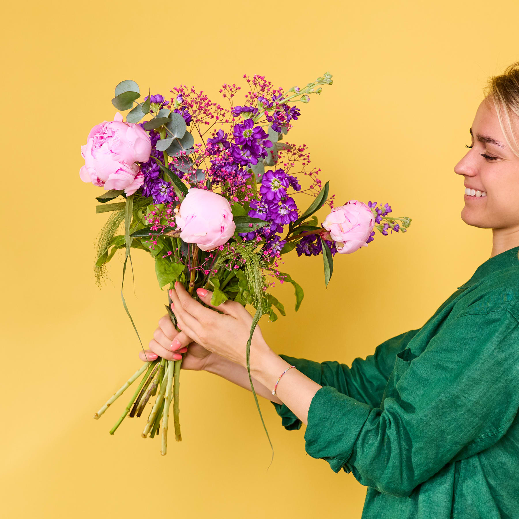 Ein farbenfroher Strauß rosa Pfingstrosen und lila Blumen wird von einer lächelnden Person in einer grünen Bluse vor einem leuchtend gelben Hintergrund gehalten.