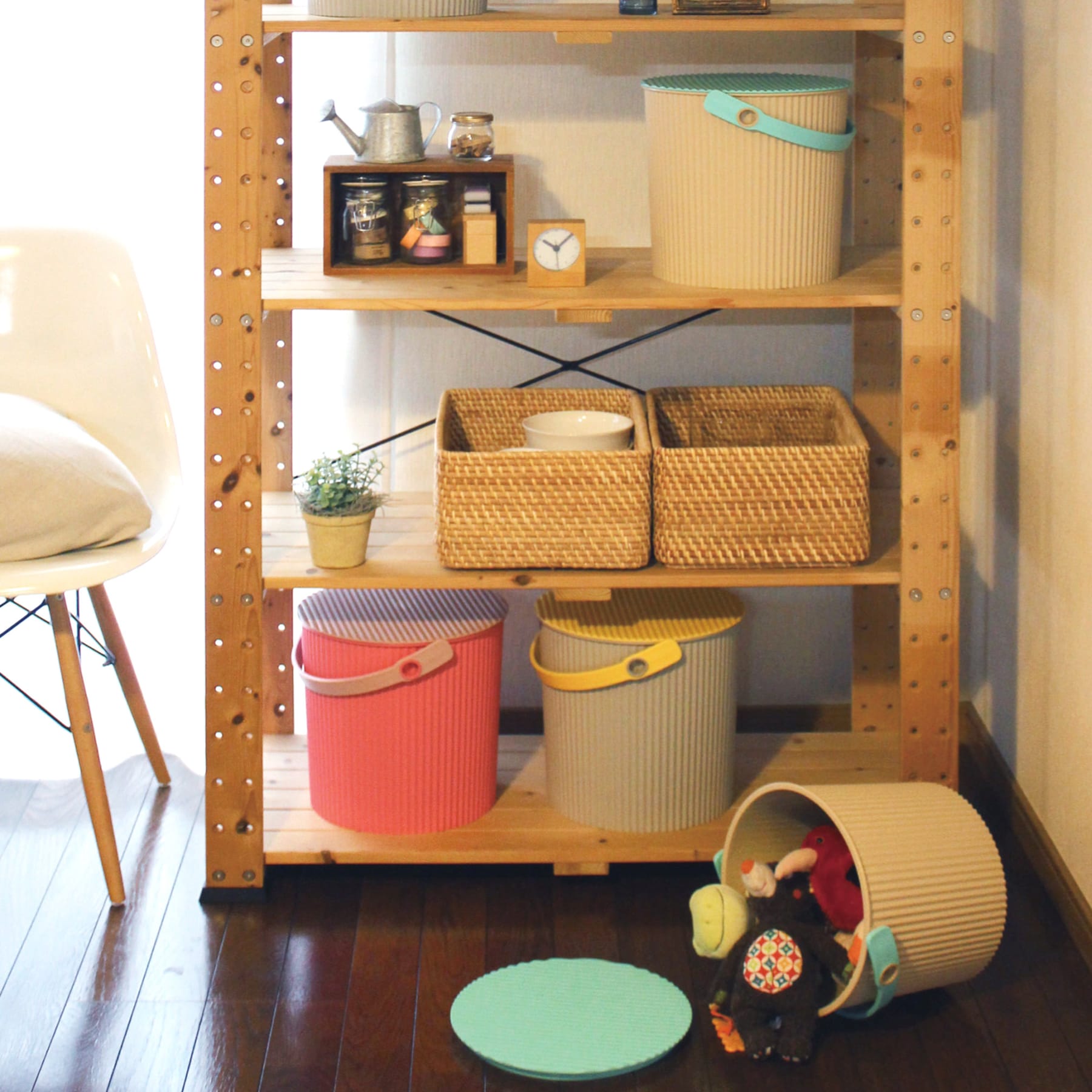 Ein Holzregal voller verschiedener Aufbewahrungsbehälter, Körbe und Dekorationsartikel, darunter auch eine Topfpflanze, steht in einem gemütlichen, gut beleuchteten Raum. Im Hintergrund ist ein Stuhl zu sehen.