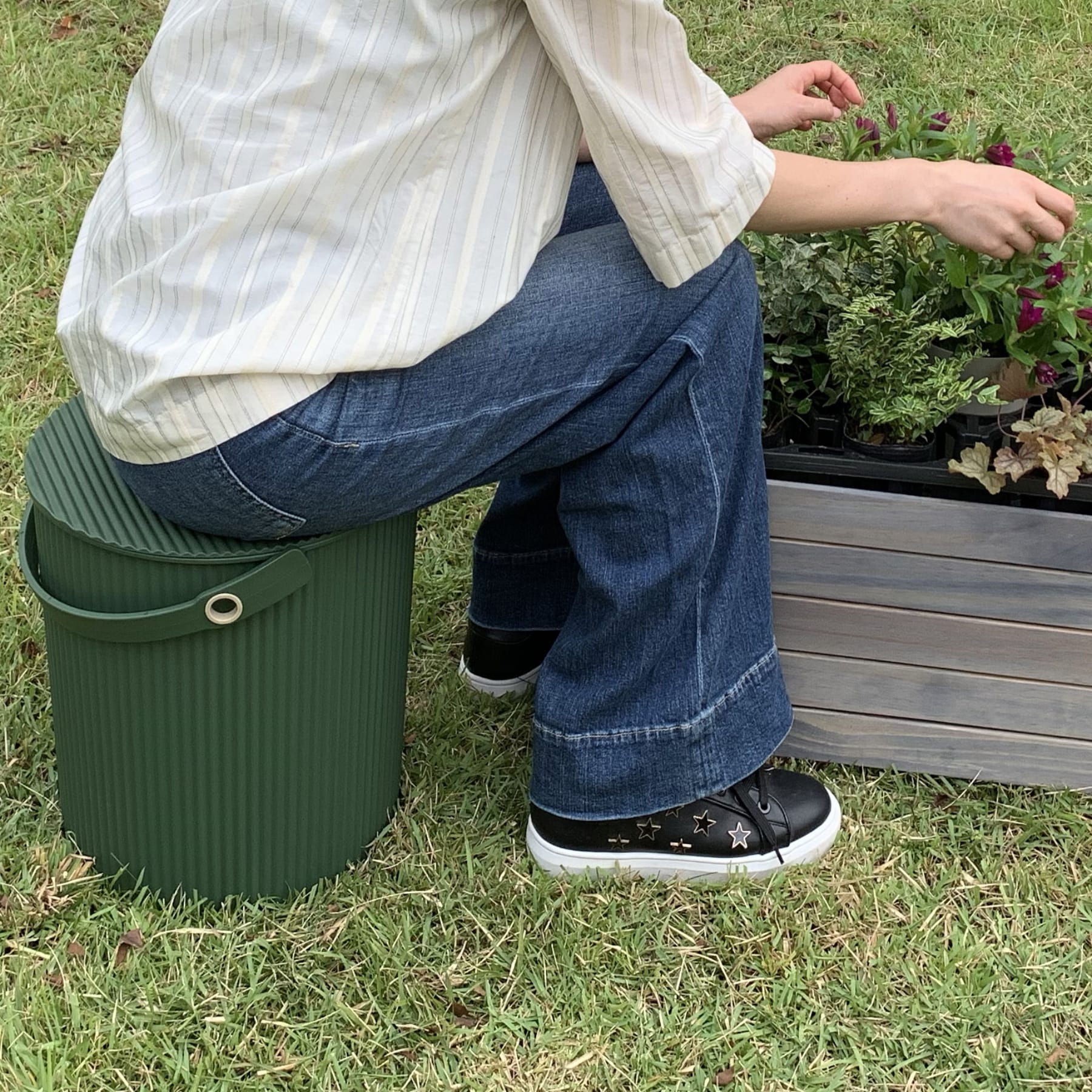 Das Bild zeigt den Unterkörper einer Person, die auf einem grünen Plastikbehälter sitzt. Sie trägt Blue Jeans und schwarz-weiße Turnschuhe, im Hintergrund ist ein Gartenbeet zu sehen.