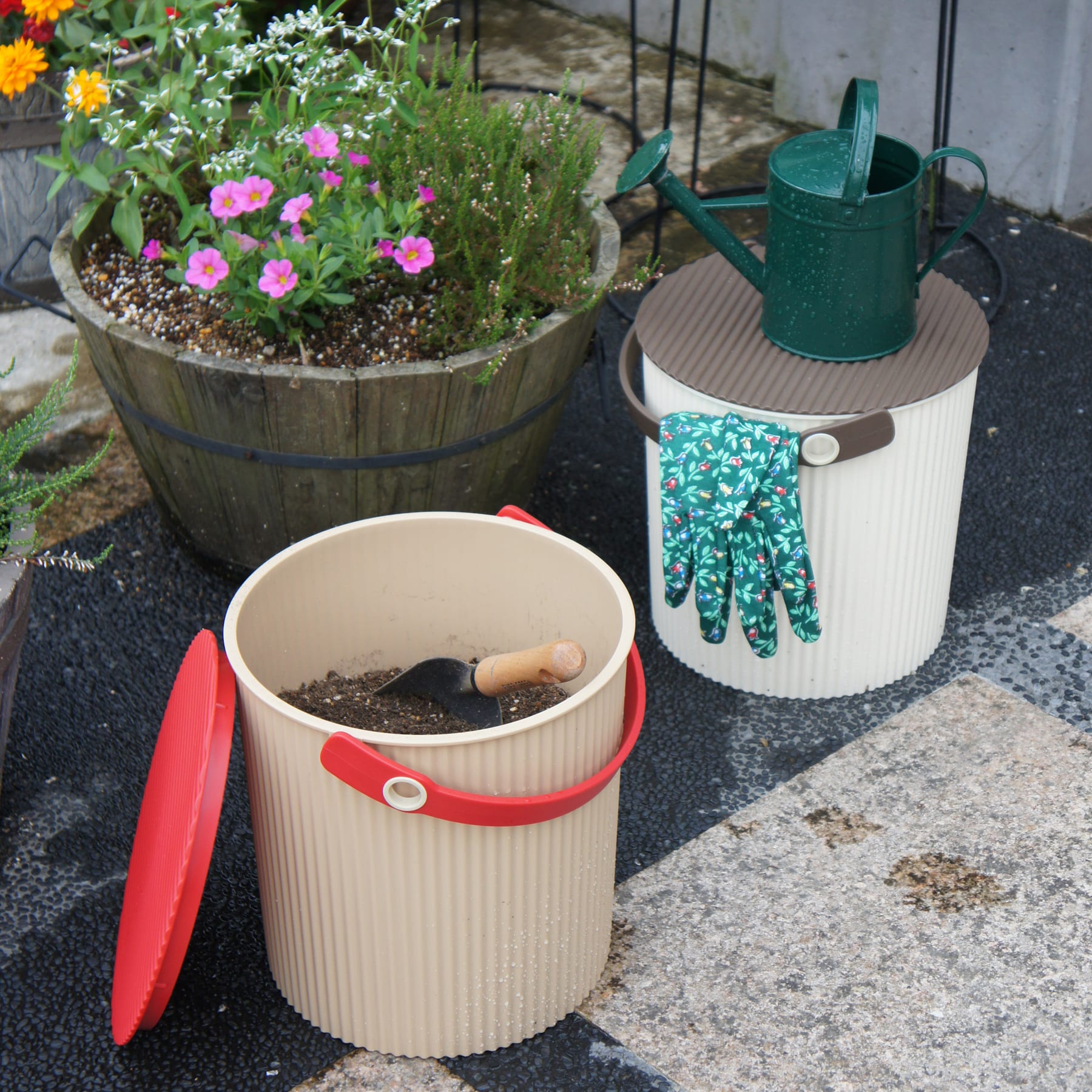 Auf einer gepflasterten Fläche sind ein bunter Blumentopf, eine Gießkanne und ein Behälter für Gartengeräte angeordnet, im Hintergrund sind ein Holzzaun und weitere Pflanzen zu sehen.