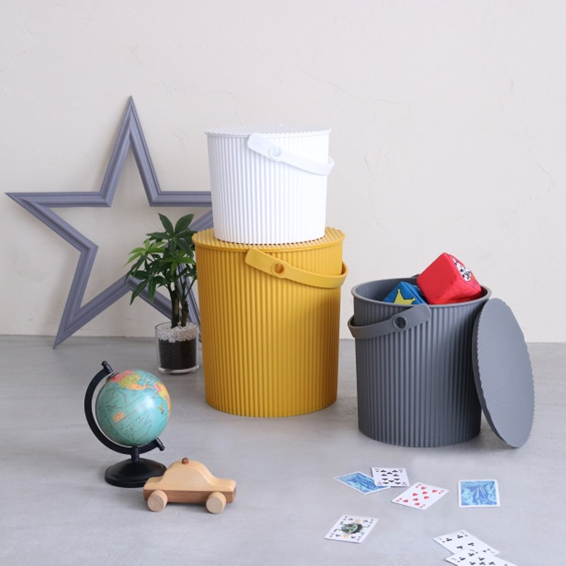 Das Bild zeigt einen minimalistischen und organisierten Arbeitsplatz mit verschiedenen dekorativen und funktionalen Elementen wie einer sternförmigen Wanddekoration, einer Topfpflanze, einem Globus, einem hölzernen Spielzeugauto und Aufbewahrungsbehältern in unterschiedlichen Farben und Texturen.