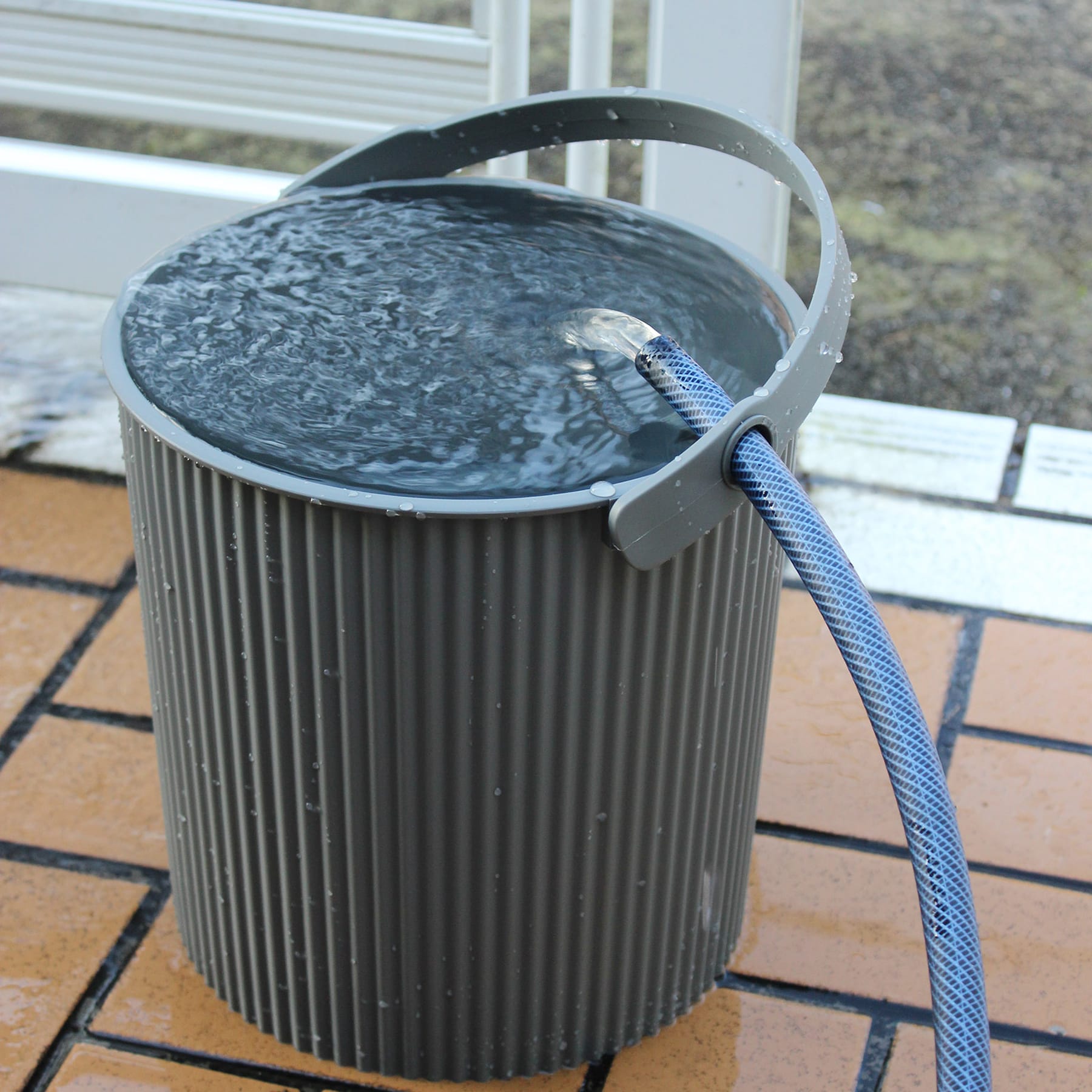 Ein schwarzer zylindrischer Behälter mit einem daran befestigten blauen Seil steht auf einer gefliesten Außenfläche, vor dem unscharfen Hintergrund, der wie ein Gebäude oder eine Struktur aussieht.