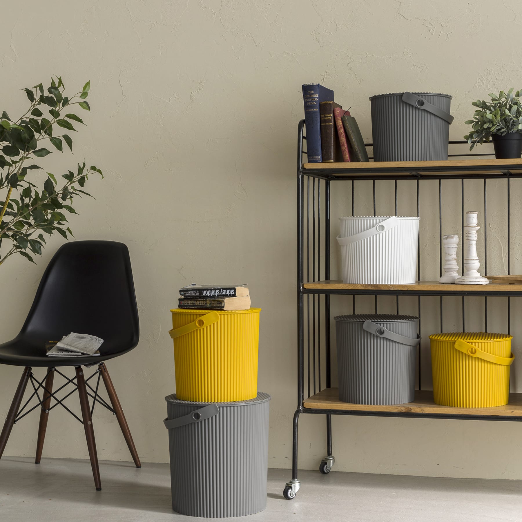 Ein gemütliches Home-Office-Setup mit einem schwarzen Stuhl, einem Holzregal voller Bücher und Dekoration sowie verschiedenen Aufbewahrungsbehältern in Gelb- und Grautönen.