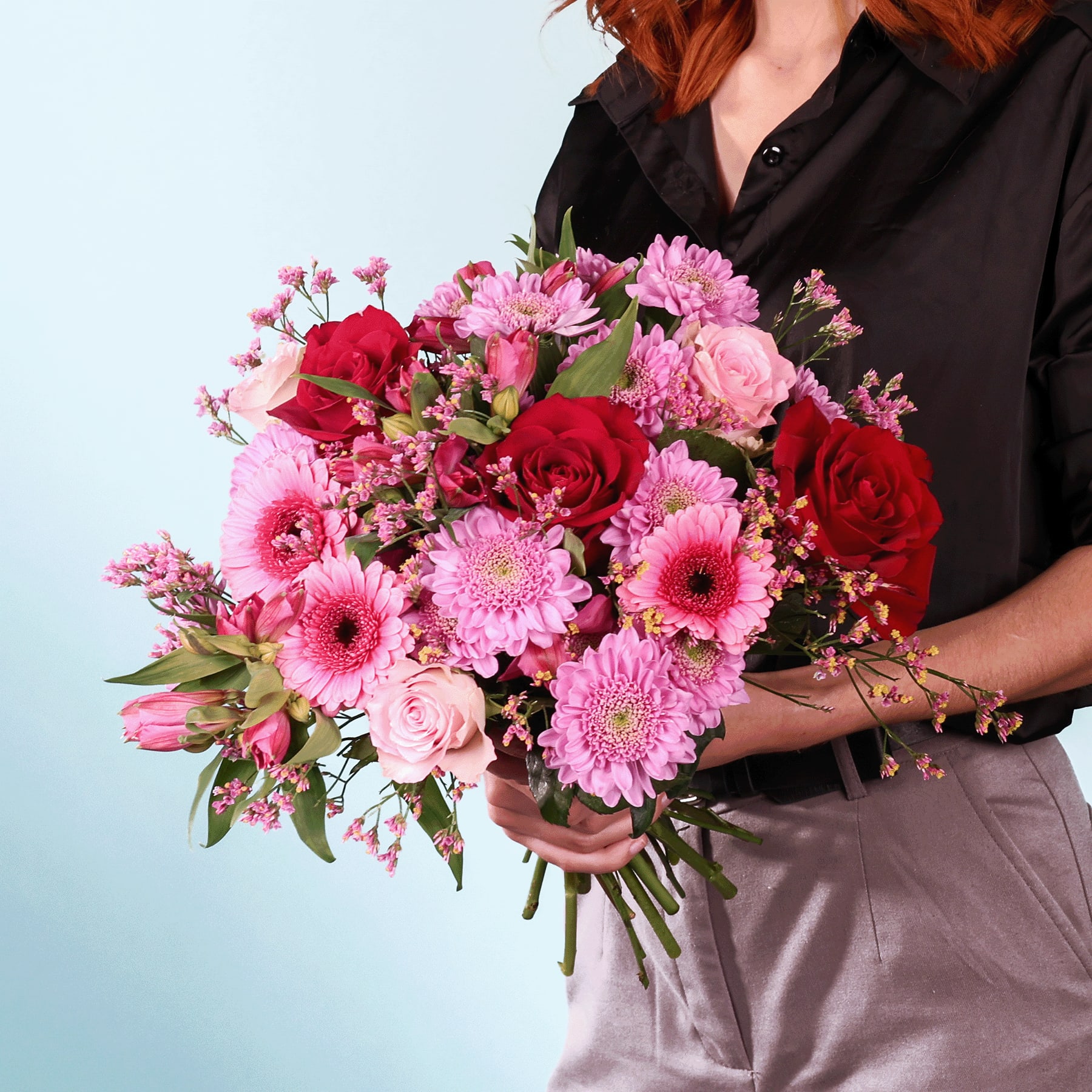 Ein leuchtender Strauß roter Rosen, rosa Gerbera und anderer bunter Blumen wird von einer Person in einem schwarzen Hemd vor einem schlichten Hintergrund gehalten.
