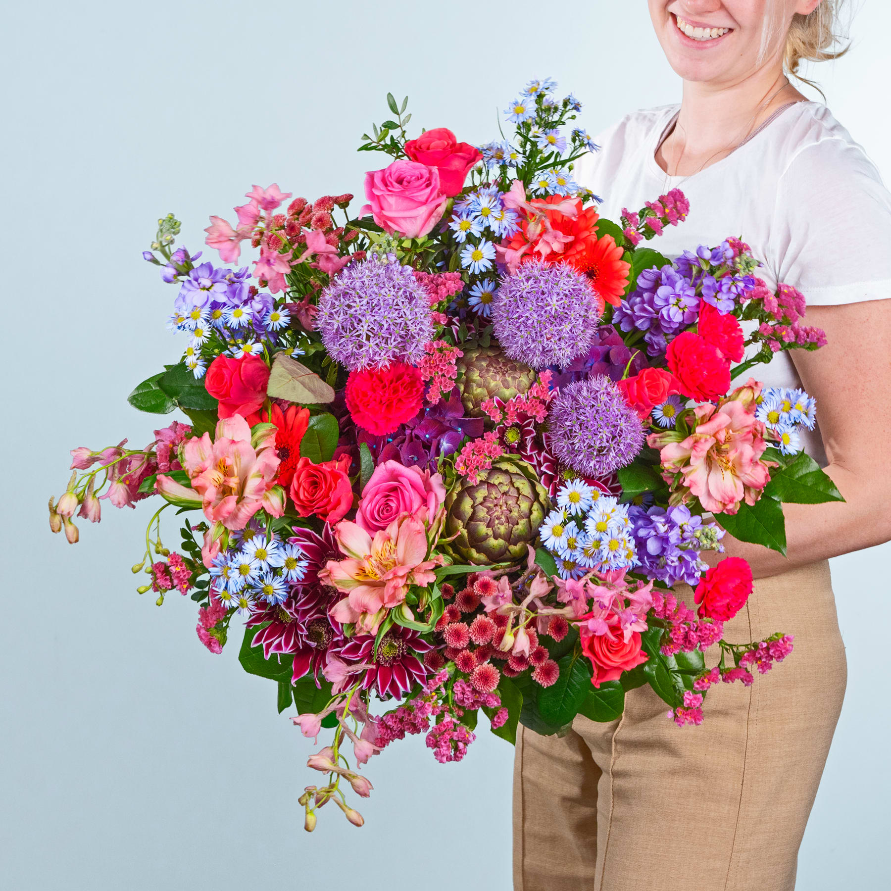 Ein leuchtender Strauß bunter Blumen, darunter Rosen, Dahlien und Allium, wird von einer lächelnden Frau in einem weißen Hemd vor einem schlichten Hintergrund gehalten.