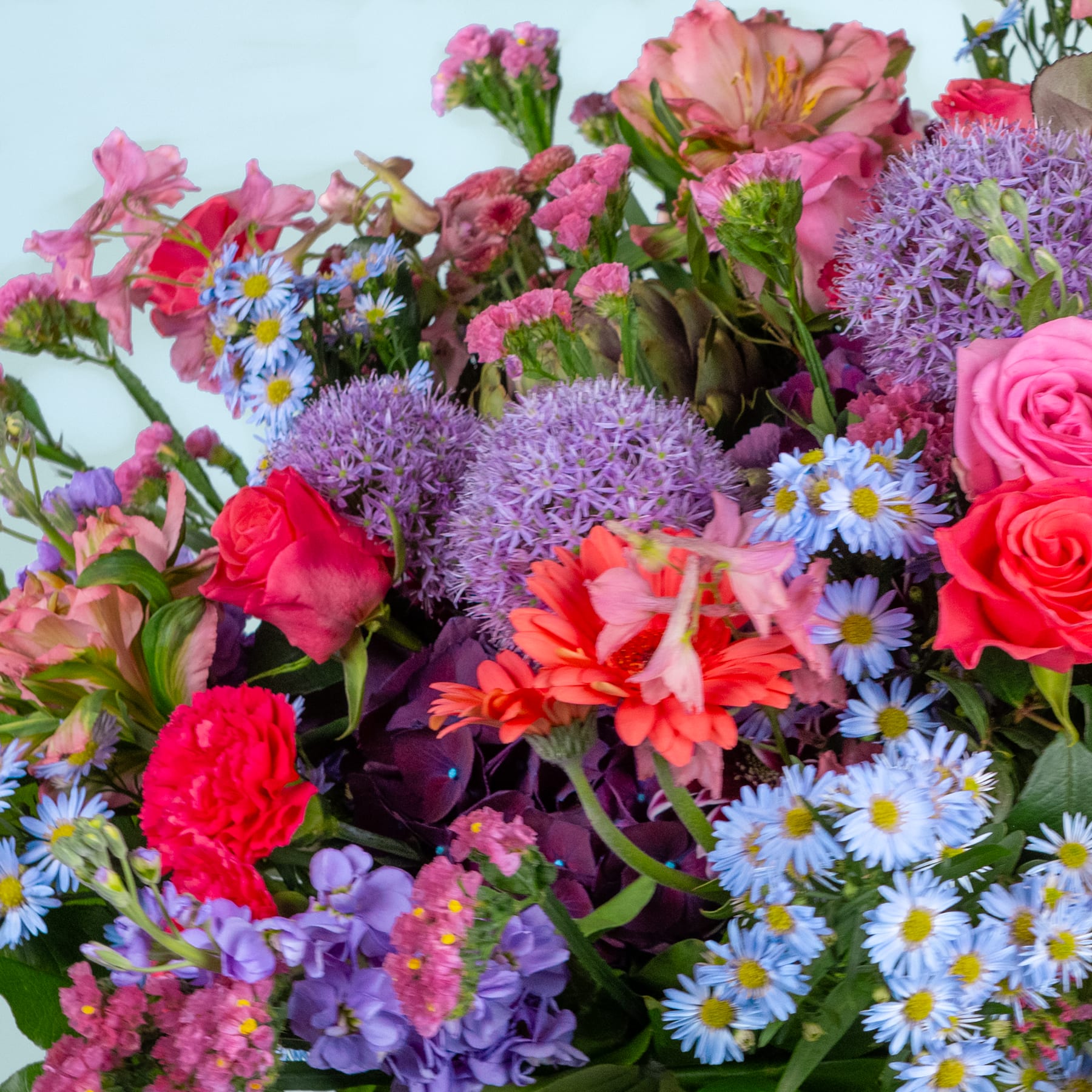Ein lebendiger Strauß verschiedener bunter Blumen, darunter Rosen, Gänseblümchen und lila Allium, vor einem sanften blauen Hintergrund.