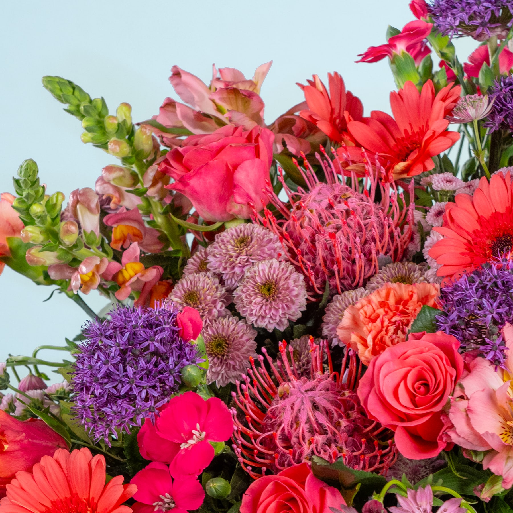 Ein lebendiger Strauß verschiedener bunter Blumen, darunter rote Rosen, rosa Nelken, violette Chrysanthemen und grünes Laub, vor einem schlichten Hintergrund.