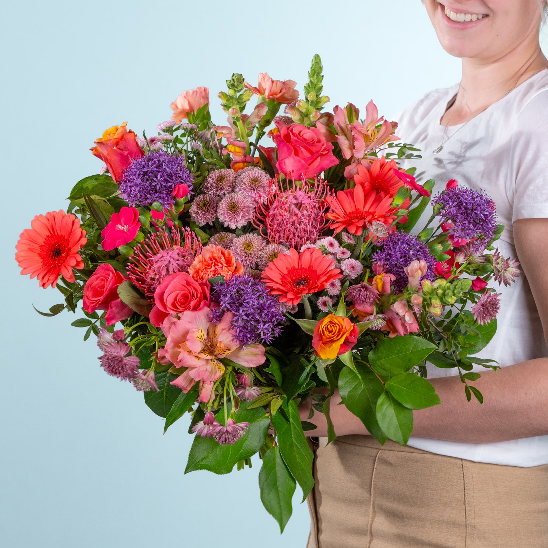 Ein lebendiger und farbenfroher Strauß verschiedener Blumen, darunter Gerbera, Rosen und Chrysanthemen, wird von einer Person im Vordergrund vor einem schlichten hellblauen Hintergrund gehalten.