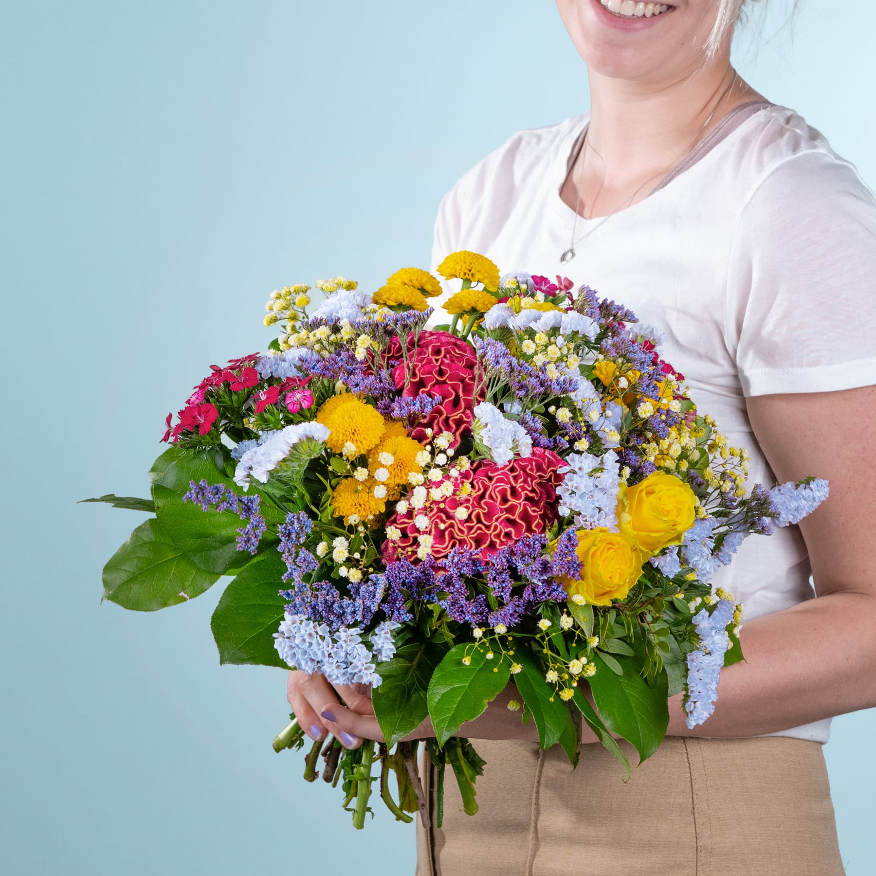 Eine lächelnde Frau hält vor einem schlichten blaugrünen Hintergrund einen leuchtend bunten Blumenstrauß, darunter rote Nelken, gelbe Chrysanthemen und violette Strandflieder.