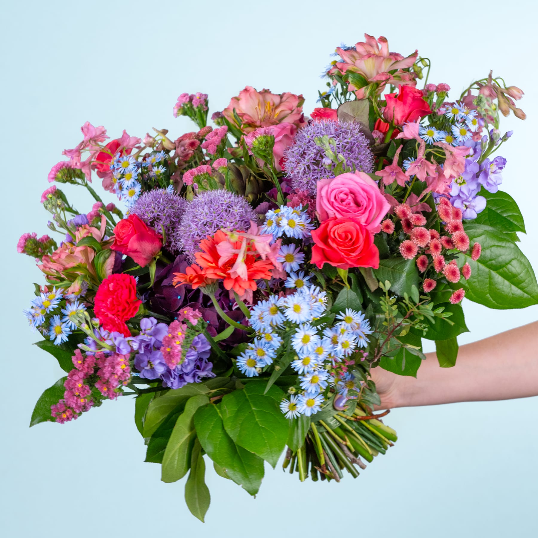 Ein leuchtender Strauß bunter Blumen, darunter Rosen, Chrysanthemen und Gänseblümchen, den eine Person vor einem schlichten blauen Hintergrund in den Händen hält.