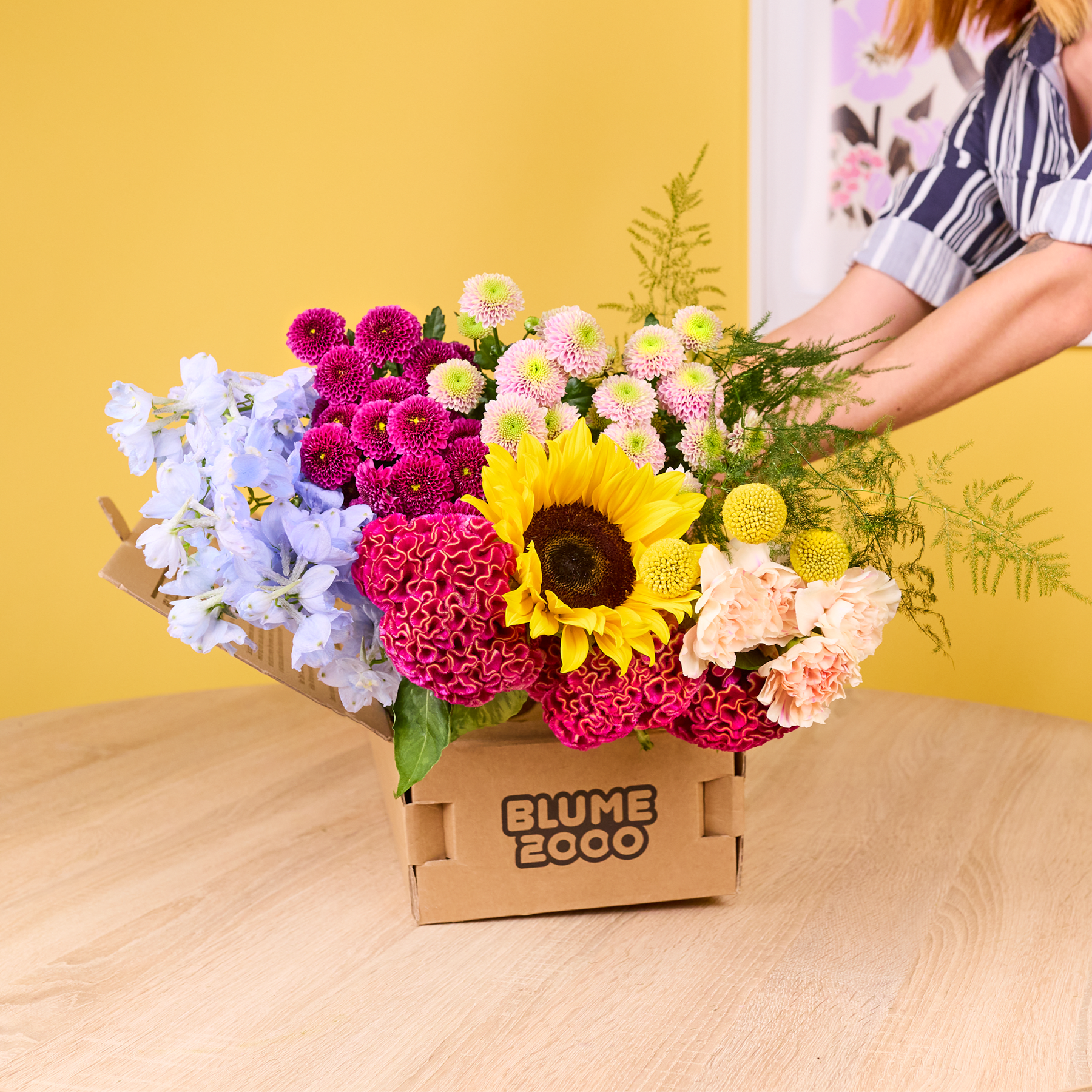 Ein lebendiger und farbenfroher Strauß verschiedener Blumen, darunter Sonnenblumen, Chrysanthemen und Dahlien, wird in einer Holzkiste auf einer Holzoberfläche vor einem gelben Hintergrund präsentiert.